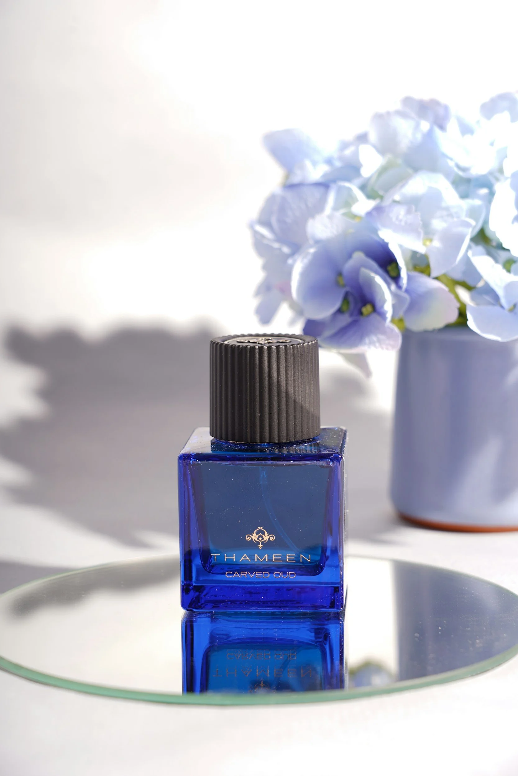 A blue bottle of Thameen Carved Oud perfume on a round mirror, with a vase of blue hydrangea flowers in the background.