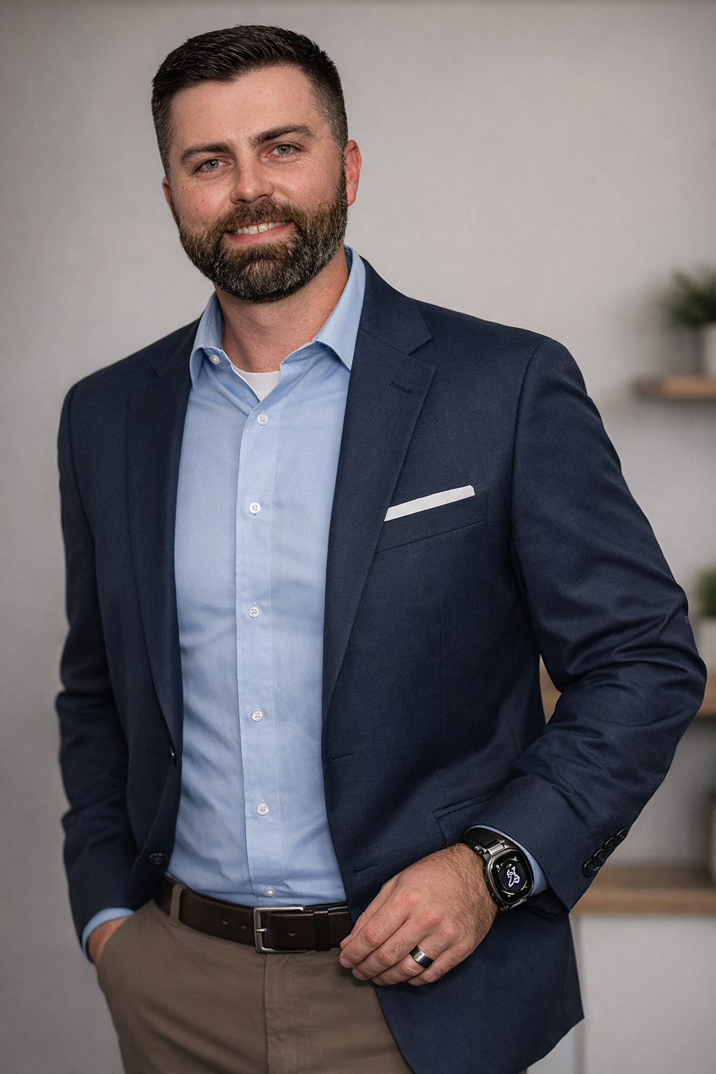 A confident man with a beard in business attire, smiling with hands in pockets, standing indoors against a plain background with a small potted plant on a shelf.