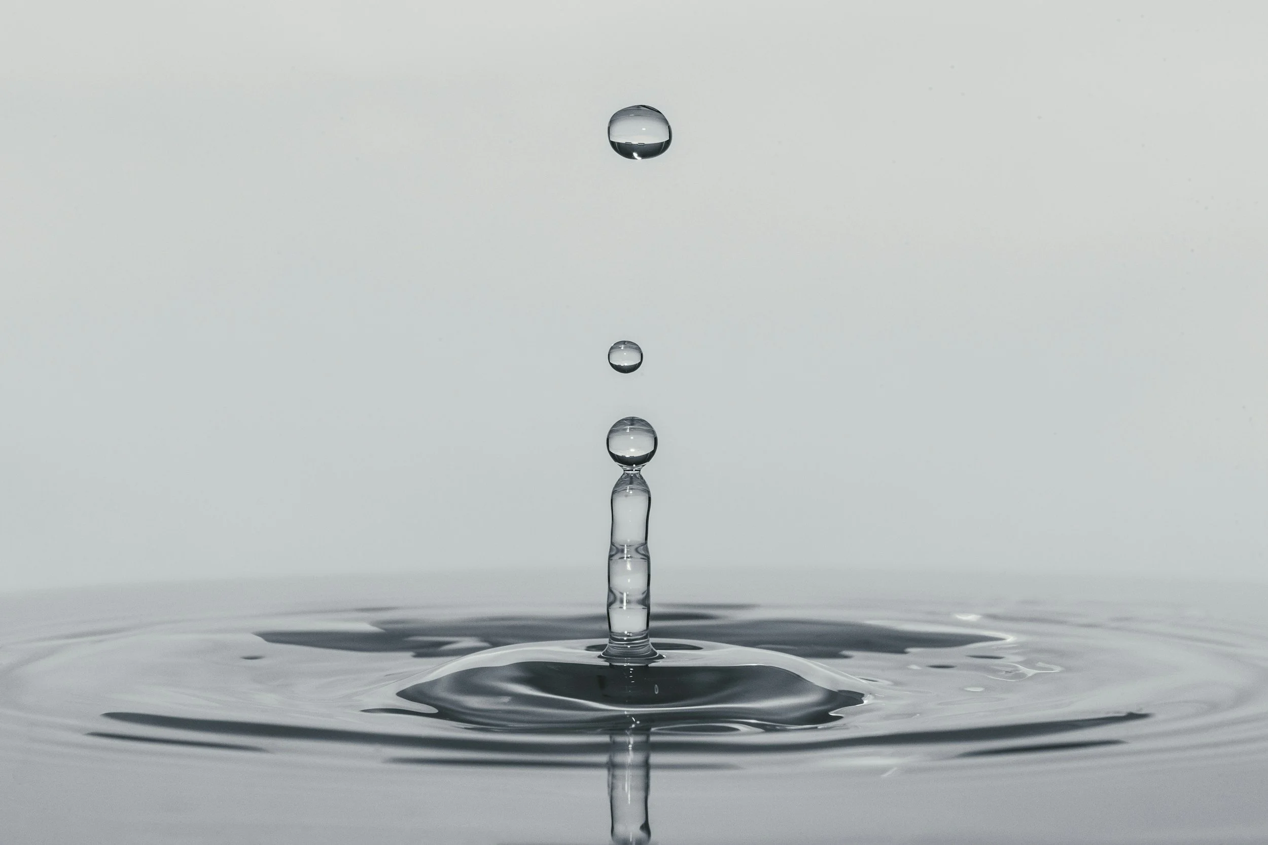 Close-up of a water droplet falling into still water, creating a splash and ripples, with a minimalist, light grey background.