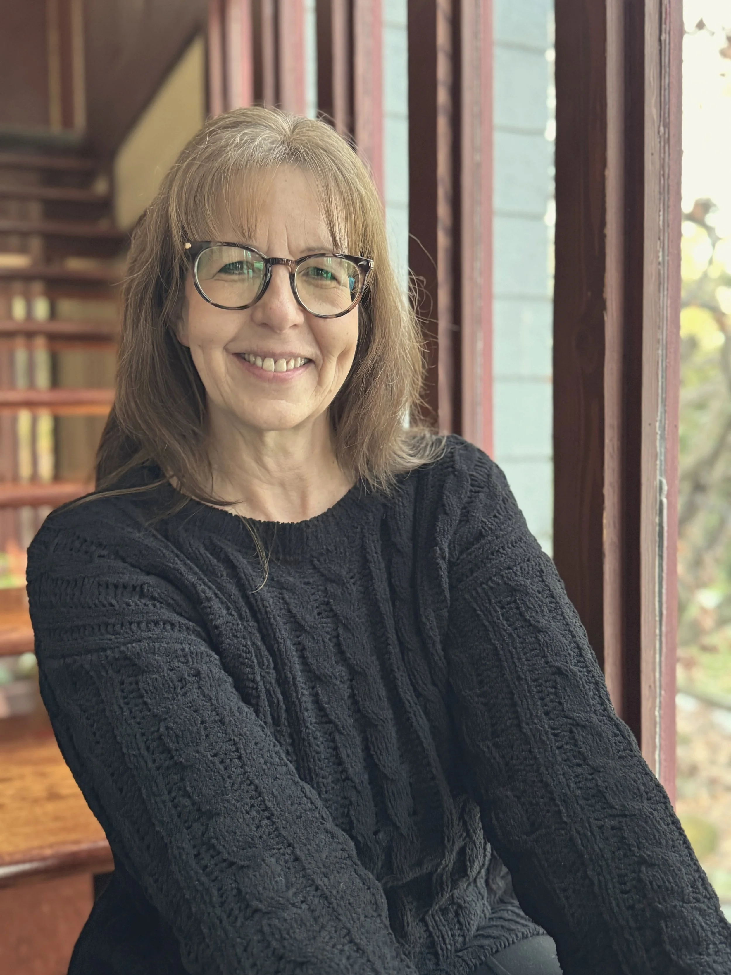 A smiling woman with shoulder-length light brown hair, wearing glasses and a black knitted sweater, sitting near a window with sunlight coming through.
