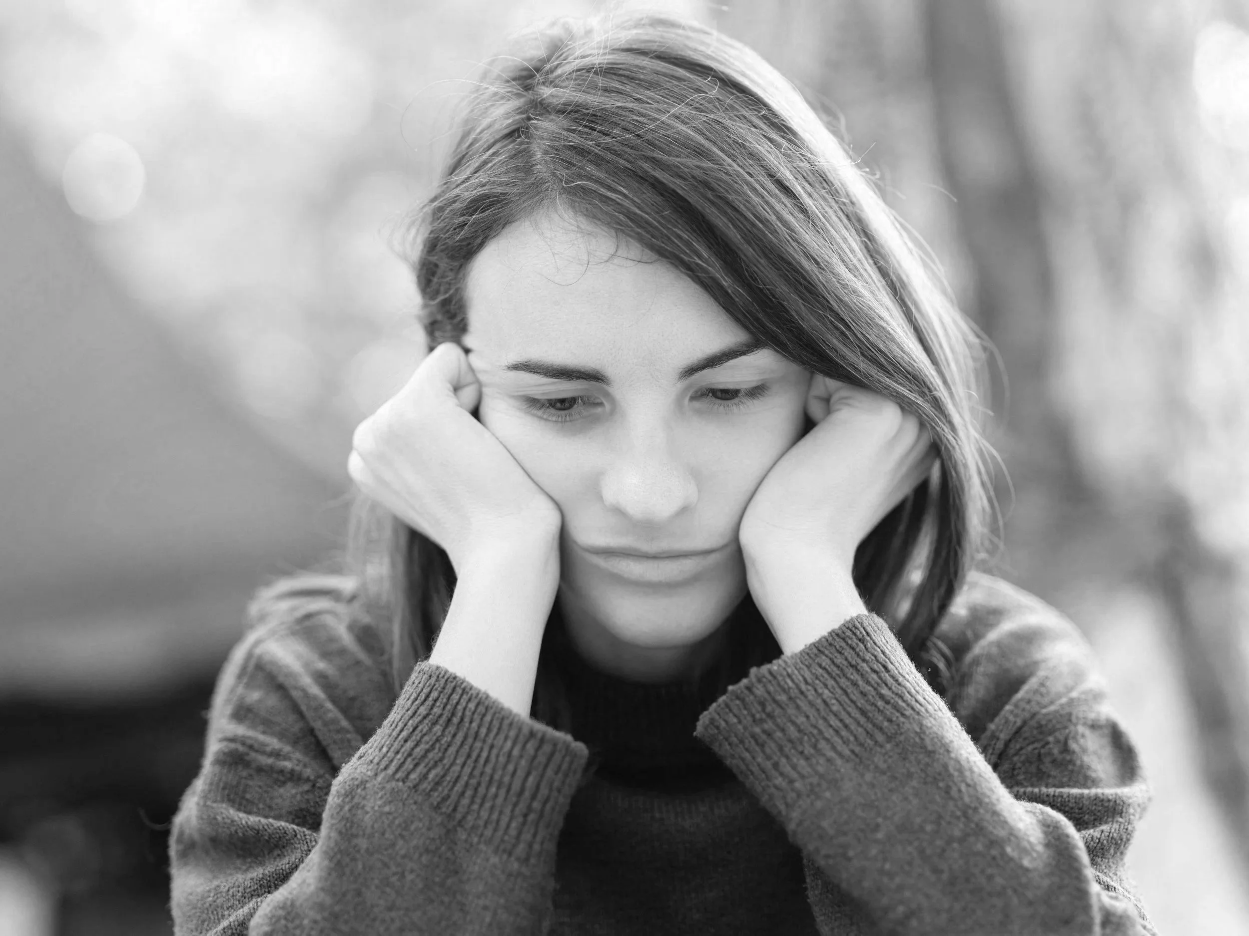 A young woman with long hair and wearing a long-sleeve sweater, resting her face in her hands outdoors with a blurred background.