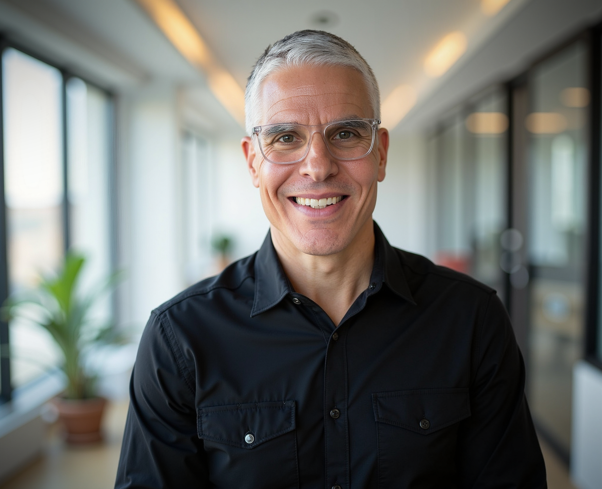 Smiling middle-aged man with short gray hair wearing clear glasses and a black shirt, in an office corridor with large windows and potted plants.