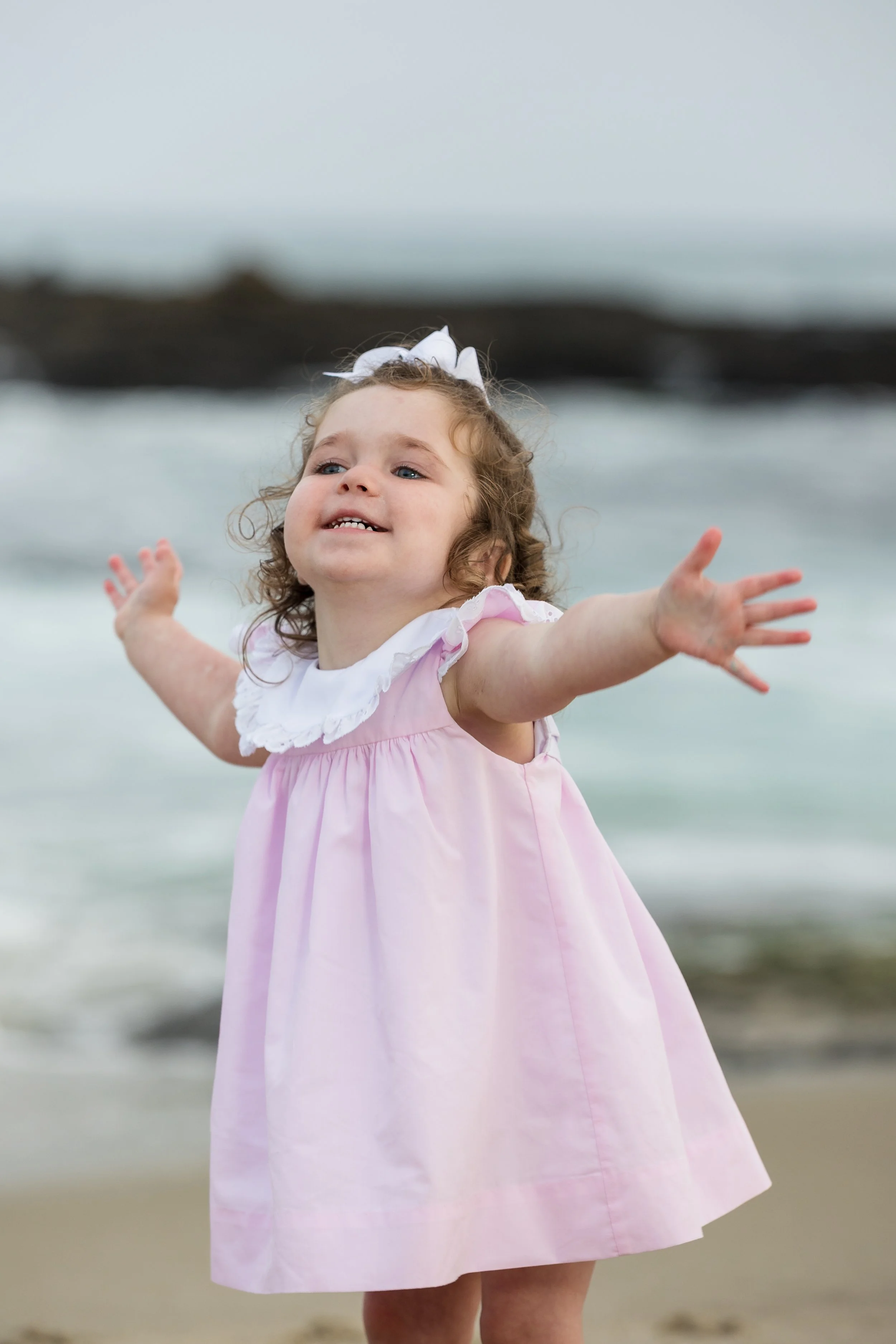 A healthy young girl with curly hair, wearing a pink dress with white lace, reaching her arms to the sky at the beach