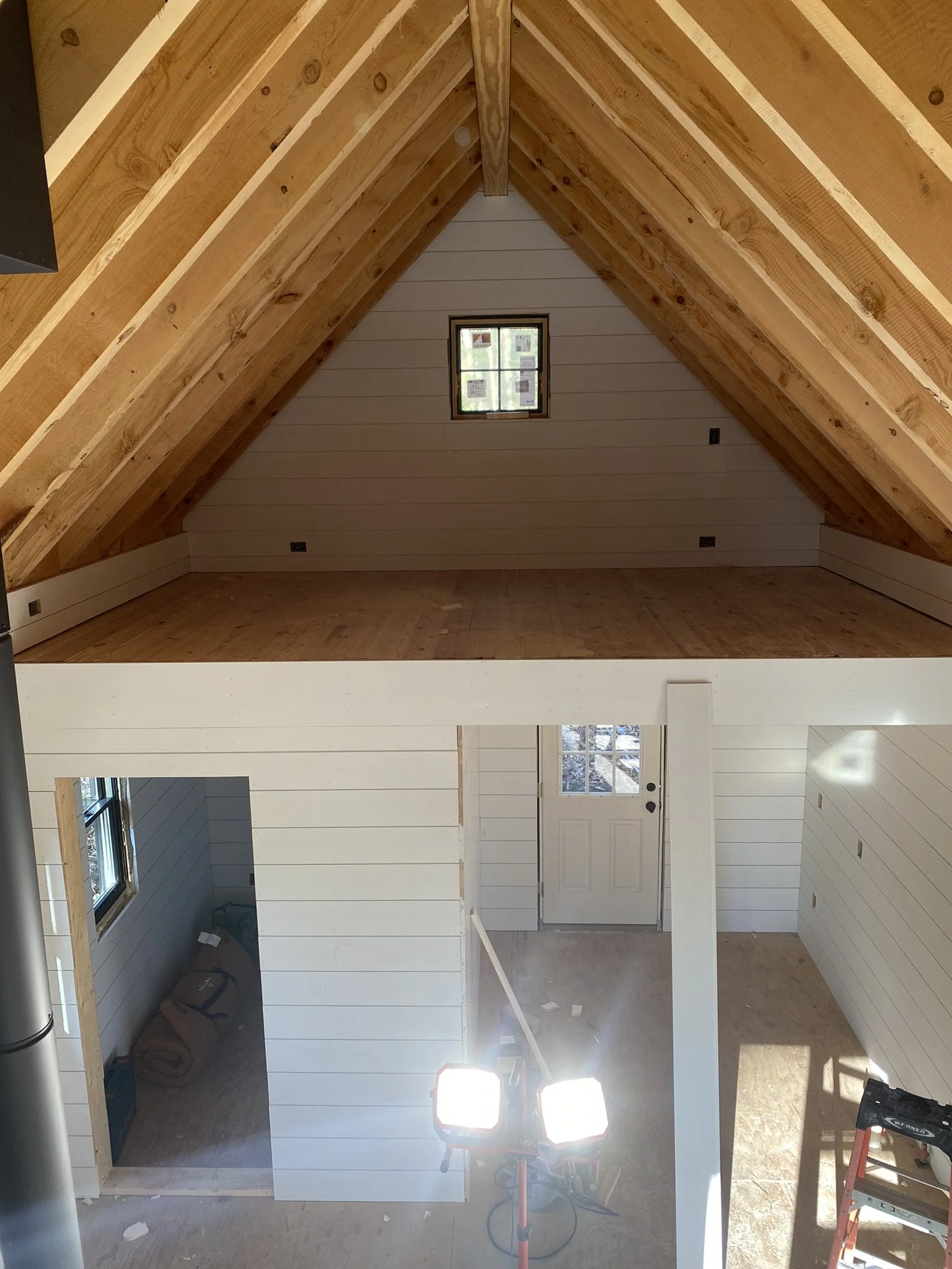 Interior of a house under construction with an open second-floor space, white shiplap walls, a small window, and construction equipment.