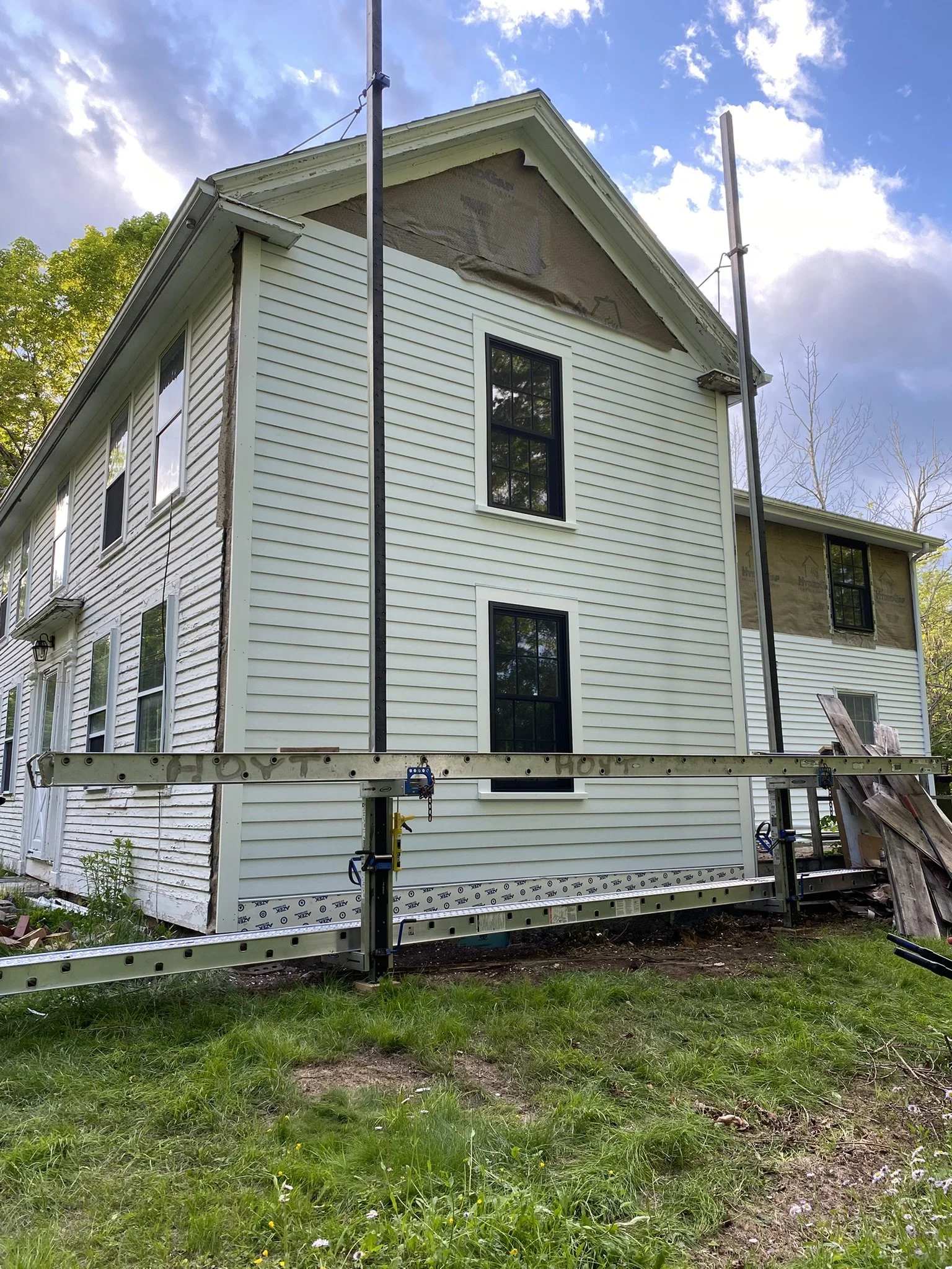 A house under renovation shows new siding being installed on the right side, with scaffolding in place. The left side shows older, weathered siding. The sky is partly cloudy with blue skies and some clouds.