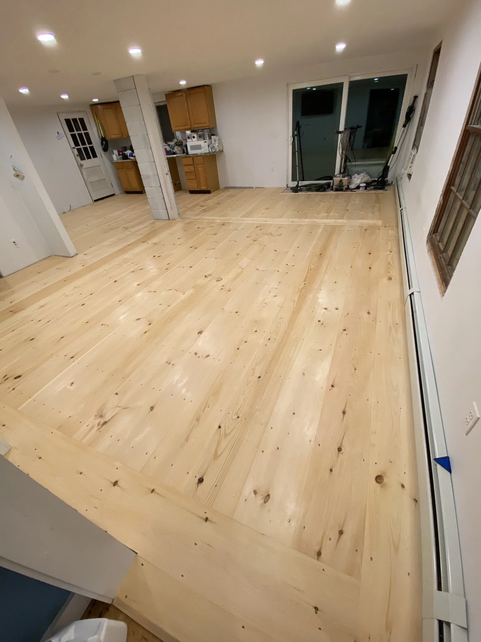 Living room with completed light-colored wooden floor, kitchen in the background, sliding glass door, and bright ceiling lights.