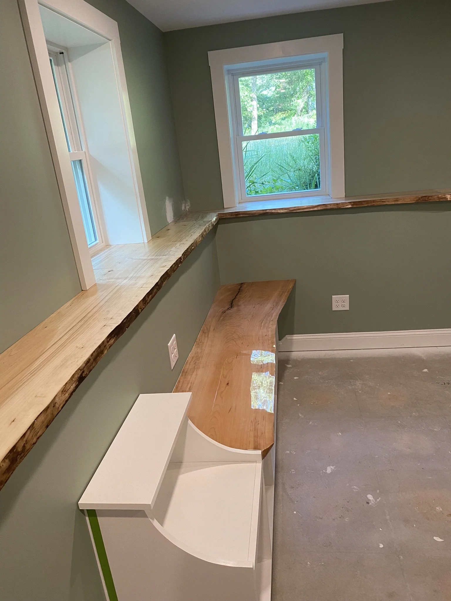 A room with green walls, a large window with white trim showing greenery outside, a built-in wooden window seat, and a smaller white piece of furniture with a curved wooden top, also positioned against the wall.