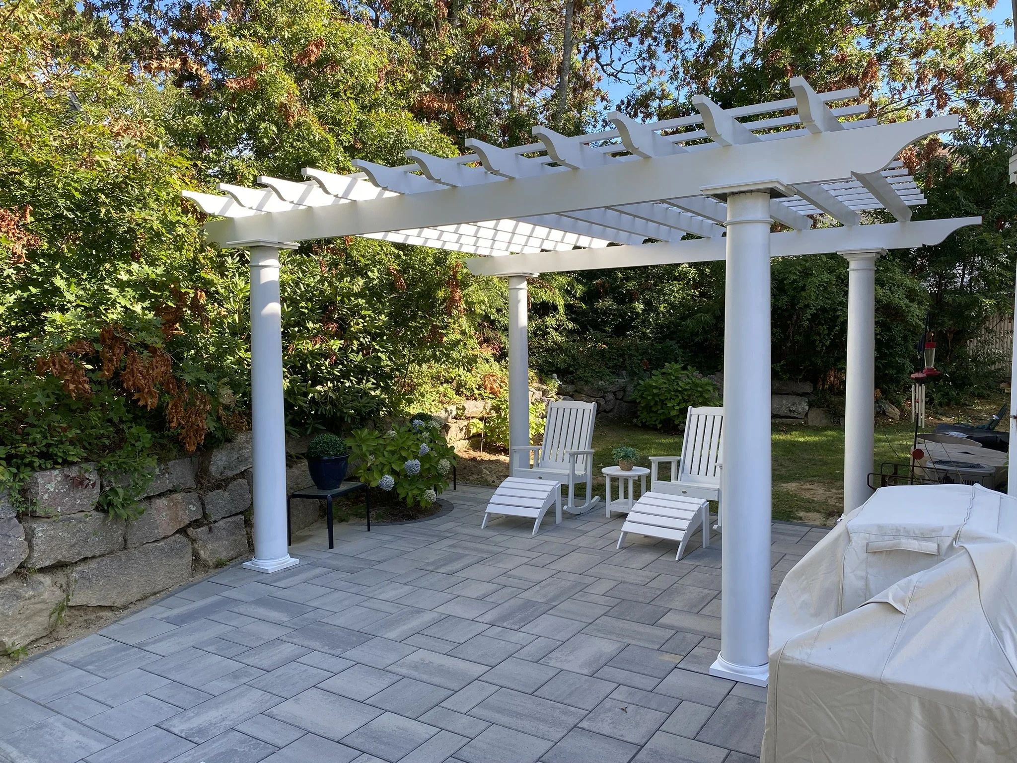 White pergola with columns over a patio area with outdoor chairs and plants in the background