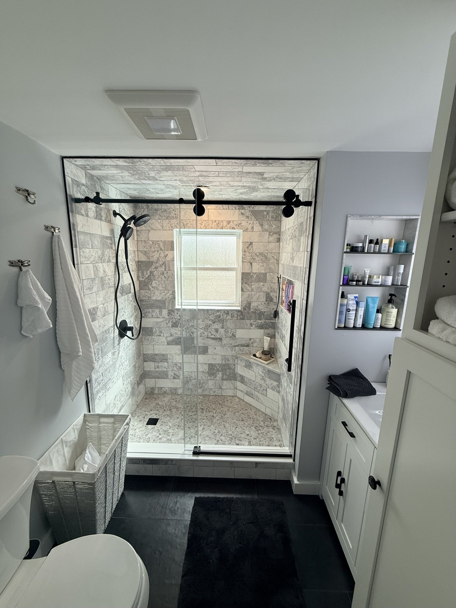 Bathroom with a walk-in shower featuring gray stone tiles, a small window, a black showerhead and fixtures, white towels hanging, a white laundry basket, a toilet, a dark rug, and a white cabinet with a black handle next to a wall shelf with toiletries.