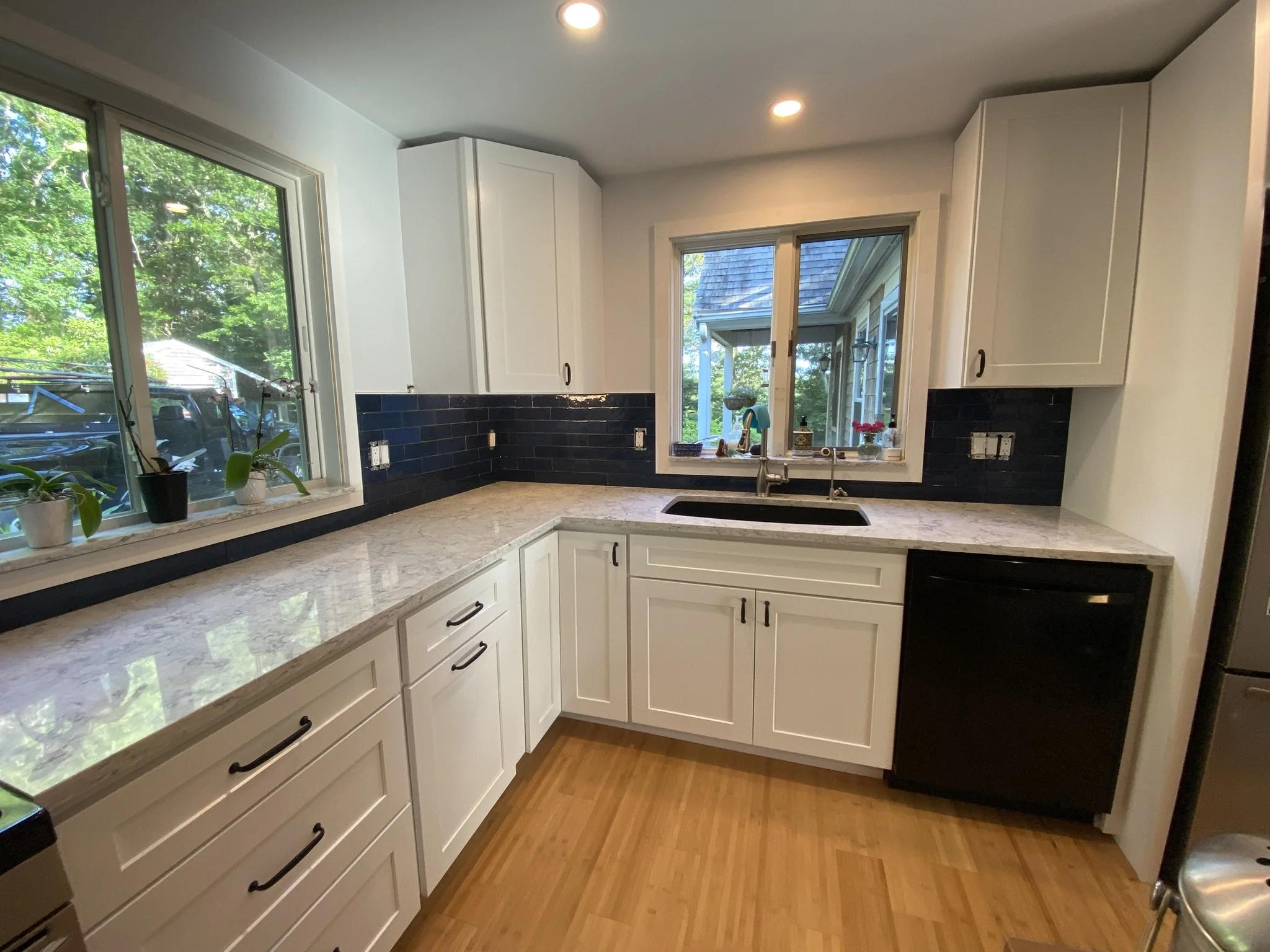 Kitchen with white cabinets, marble countertops, black backsplash, black dishwasher, two windows overlooking greenery, and potted plants on the windowsill.