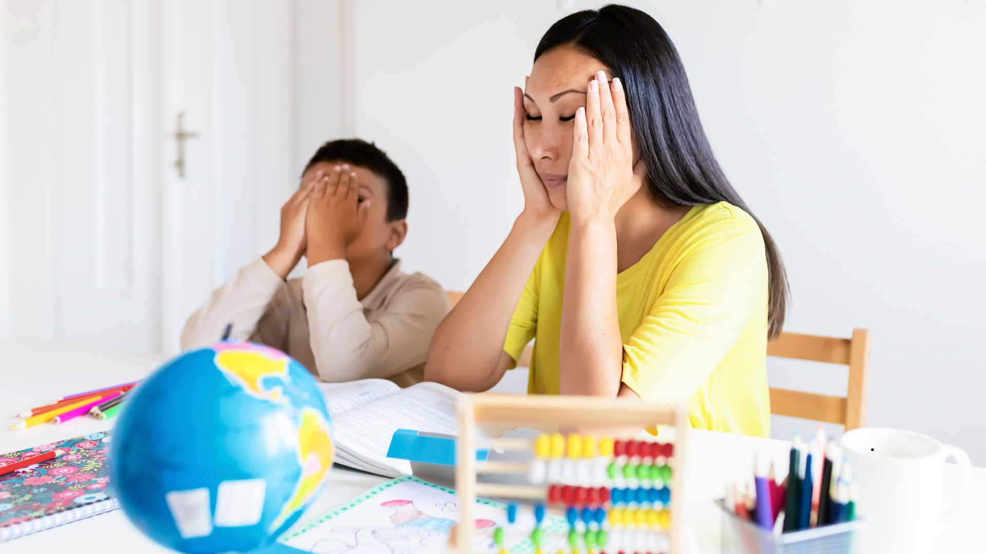A woman and a young boy sitting at a desk with their hands on their faces, appearing stressed or overwhelmed. On the desk are a globe, colorful pencils, a workbook, and school supplies.