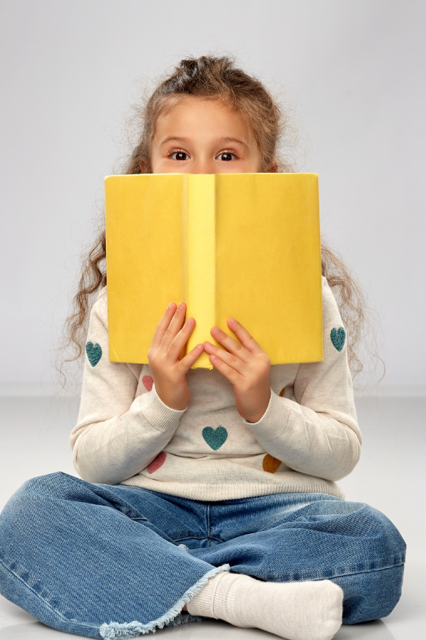 A young girl with curly hair sitting cross-legged and holding a yellow book, partially covering her face, against a plain background.