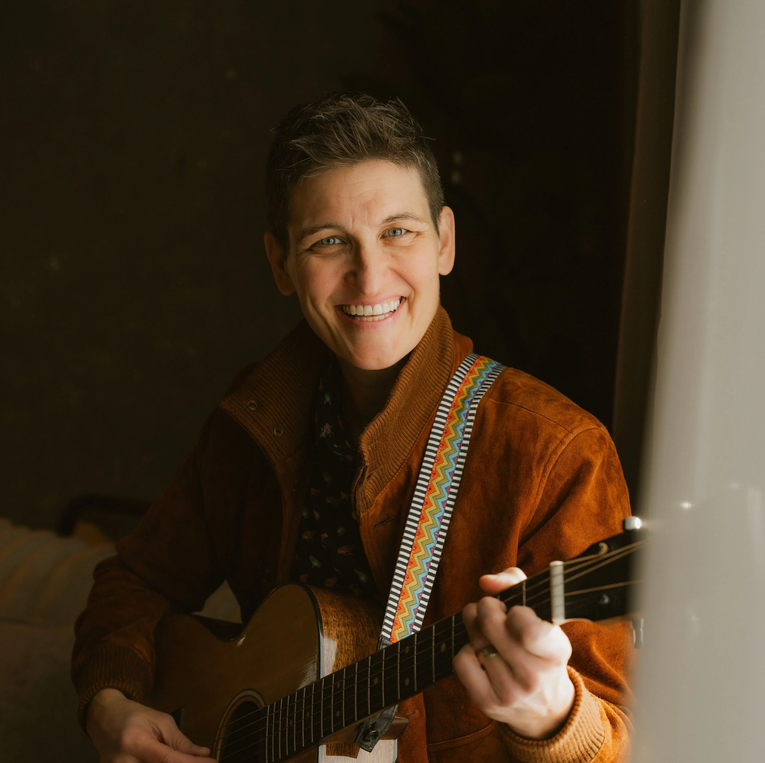 Woman with short brown hair, smiling, wearing a brown suede jacket, dark collared shirt, and holding an acoustic guitar.