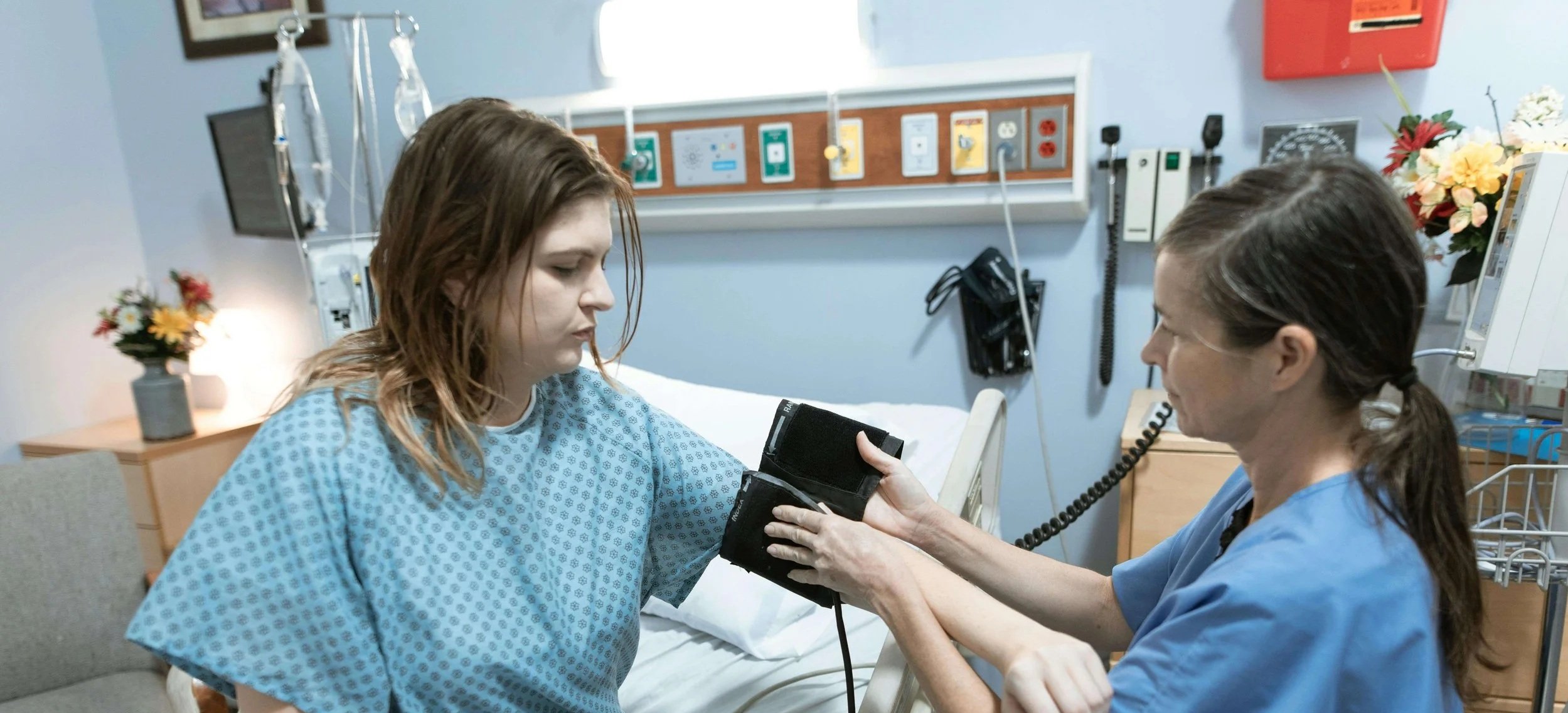 A young woman in a hospital bed during a check-up by a nurse, who is using a blood pressure cuff.