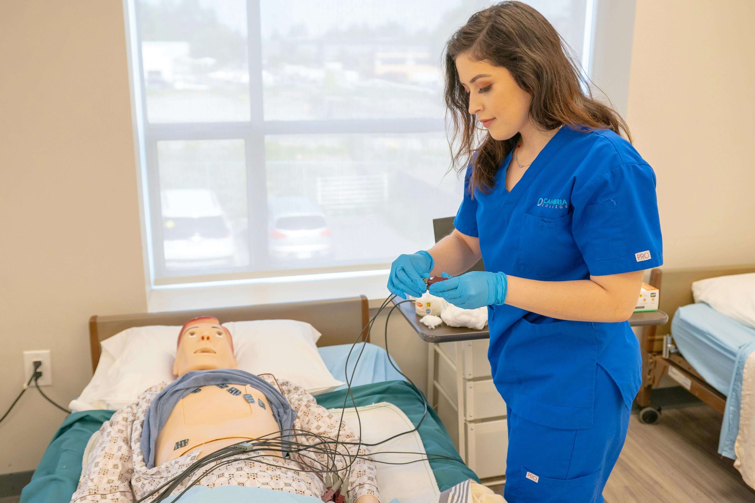 A female healthcare professional in blue scrubs and gloves tending to a medical mannequin patient on a hospital bed.
