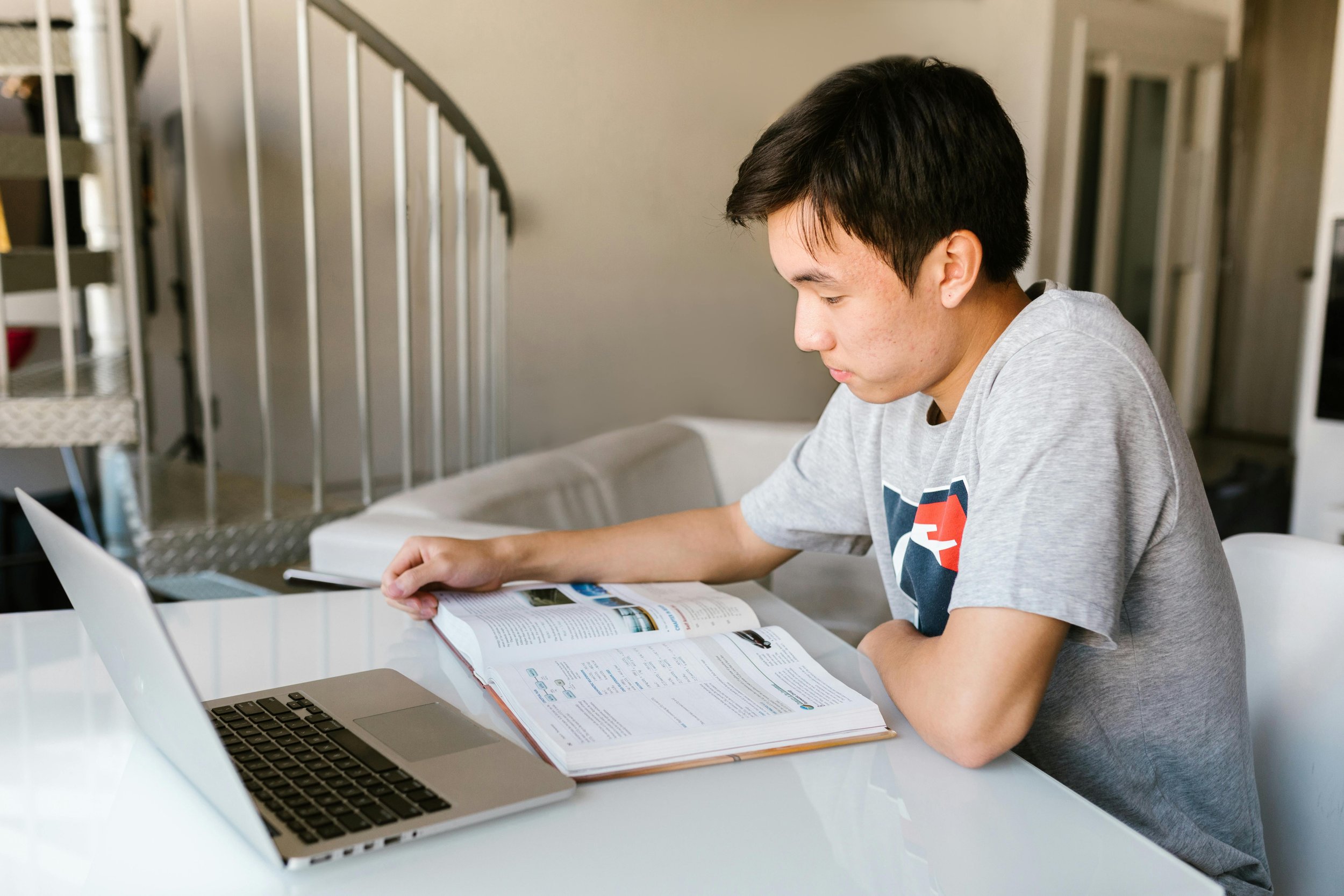 A young man with black hair wearing a gray T-shirt studies at a white desk, reading a large open book, in a modern room with a laptop, a laptop stand, and a bookshelf in the background.