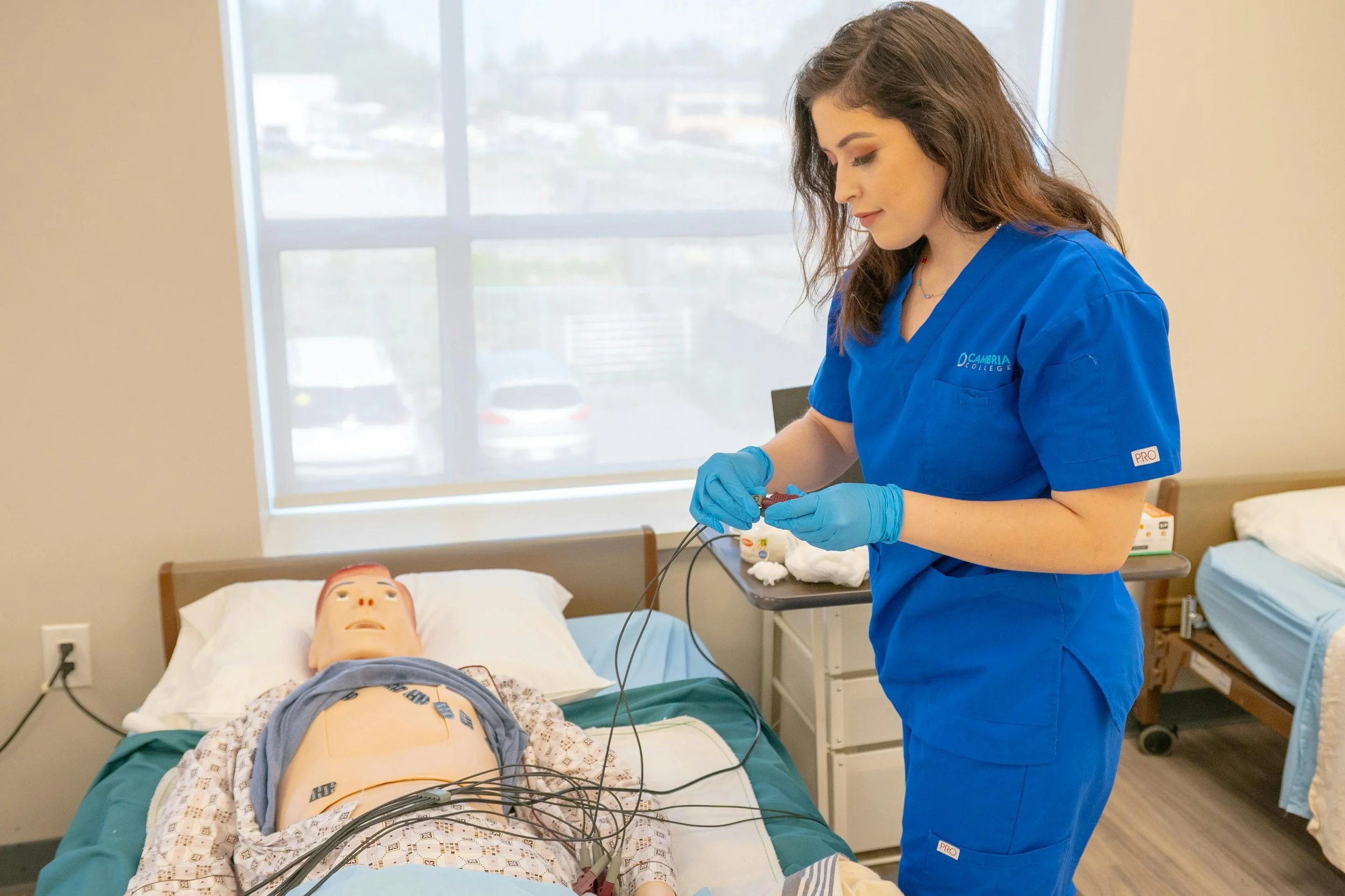 A healthcare professional in blue scrubs and gloves performs a medical procedure on a patient lying in a hospital bed with electrodes attached to their chest, in a room with a large window.