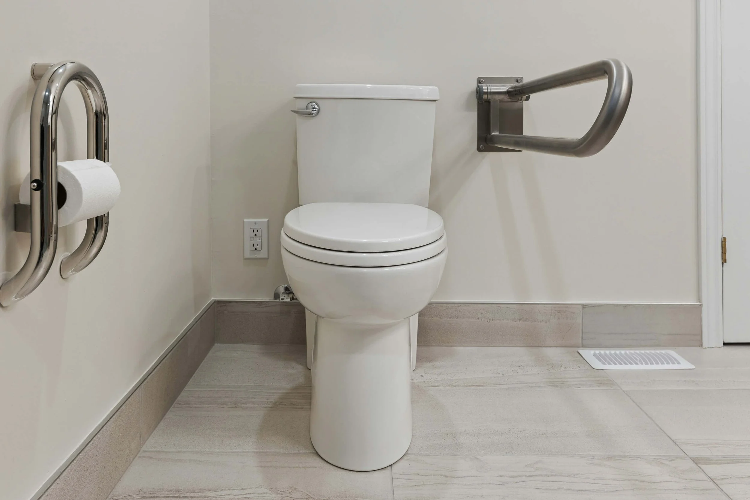 Bathroom with a toilet, fold-down grab bar, stainless steel handrails on each side, electrical outlet, and a floor vent in the corner.