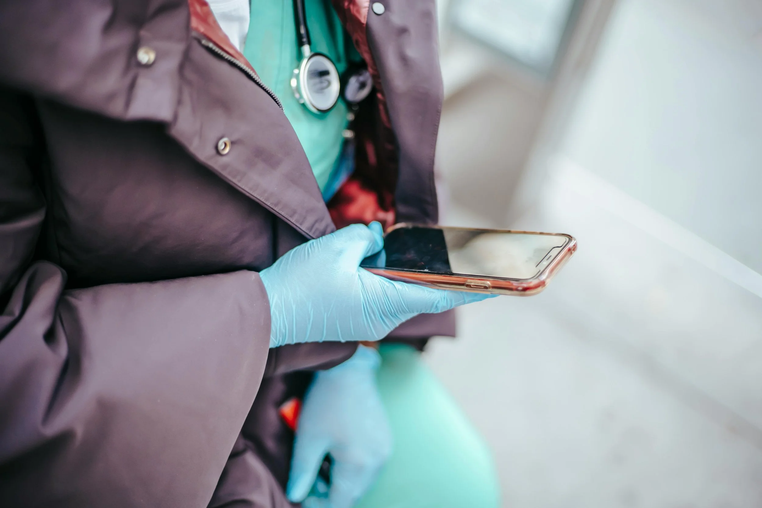 Person wearing purple gloves holding a smartphone in their hand, with a stethoscope around their neck and wearing a teal medical gown and a purple jacket, standing near a window.