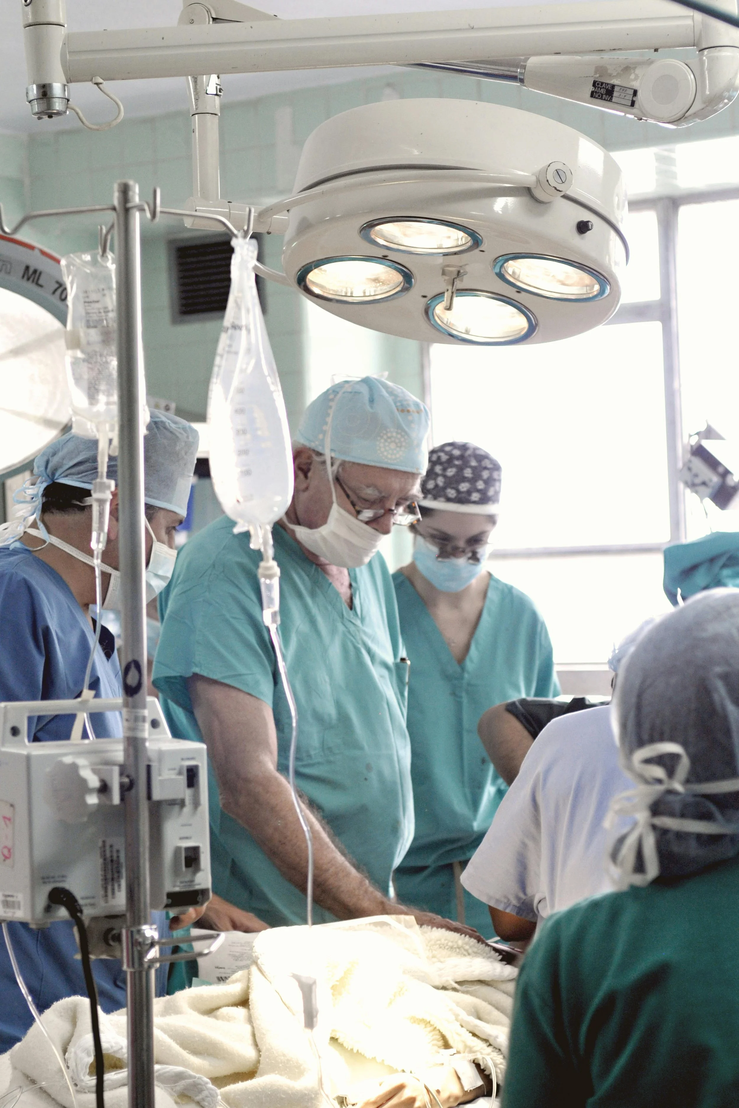 Medical team performing surgery in an operating room, with a patient covered in a blanket and surrounded by medical equipment, nurses, and doctors wearing scrubs, masks, and surgical caps.