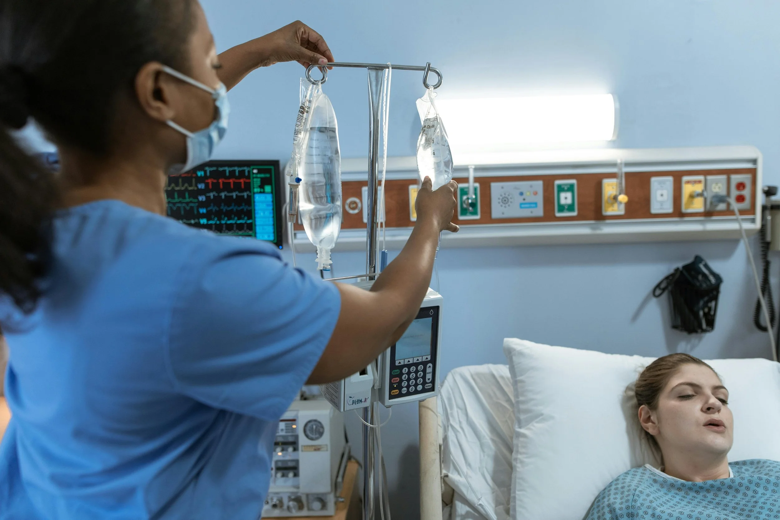 A healthcare professional in scrubs and a face mask adjusting IV fluids in a hospital room, with a patient lying in bed and monitoring equipment in the background.