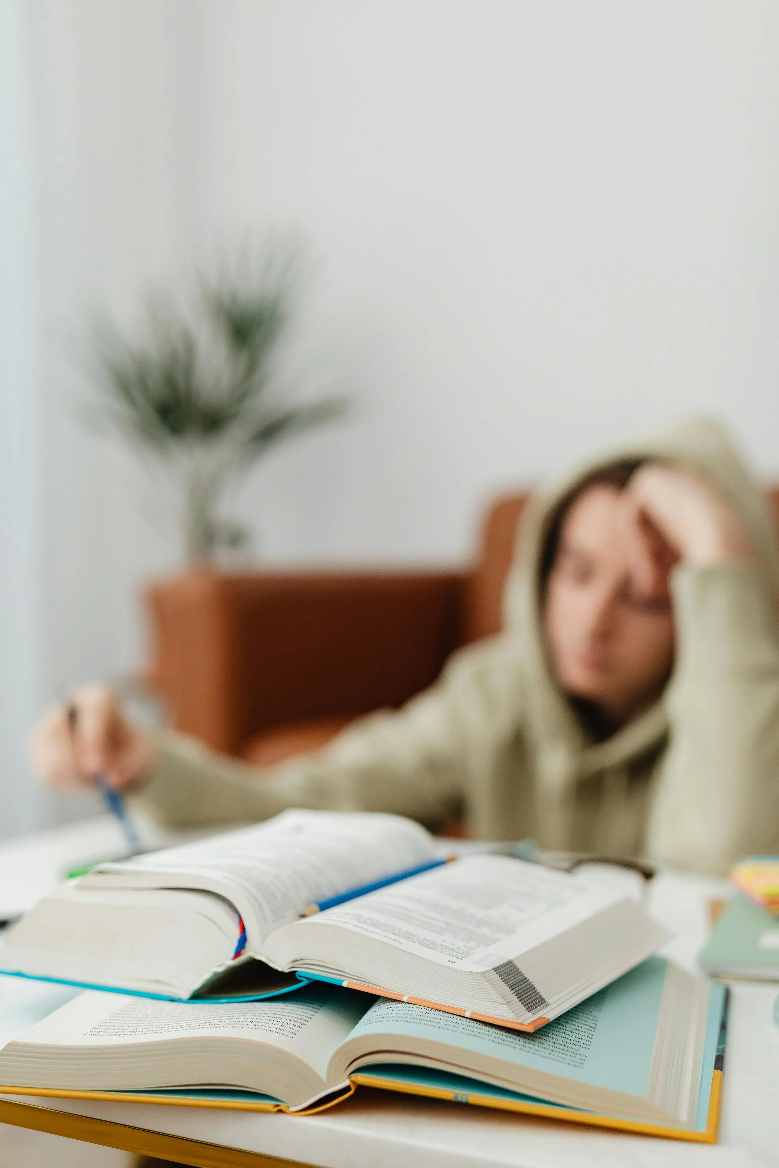 A person wearing a beige hoodie sitting at a table with open books and writing tools, studying or working. The background features a plain wall with a potted plant.
