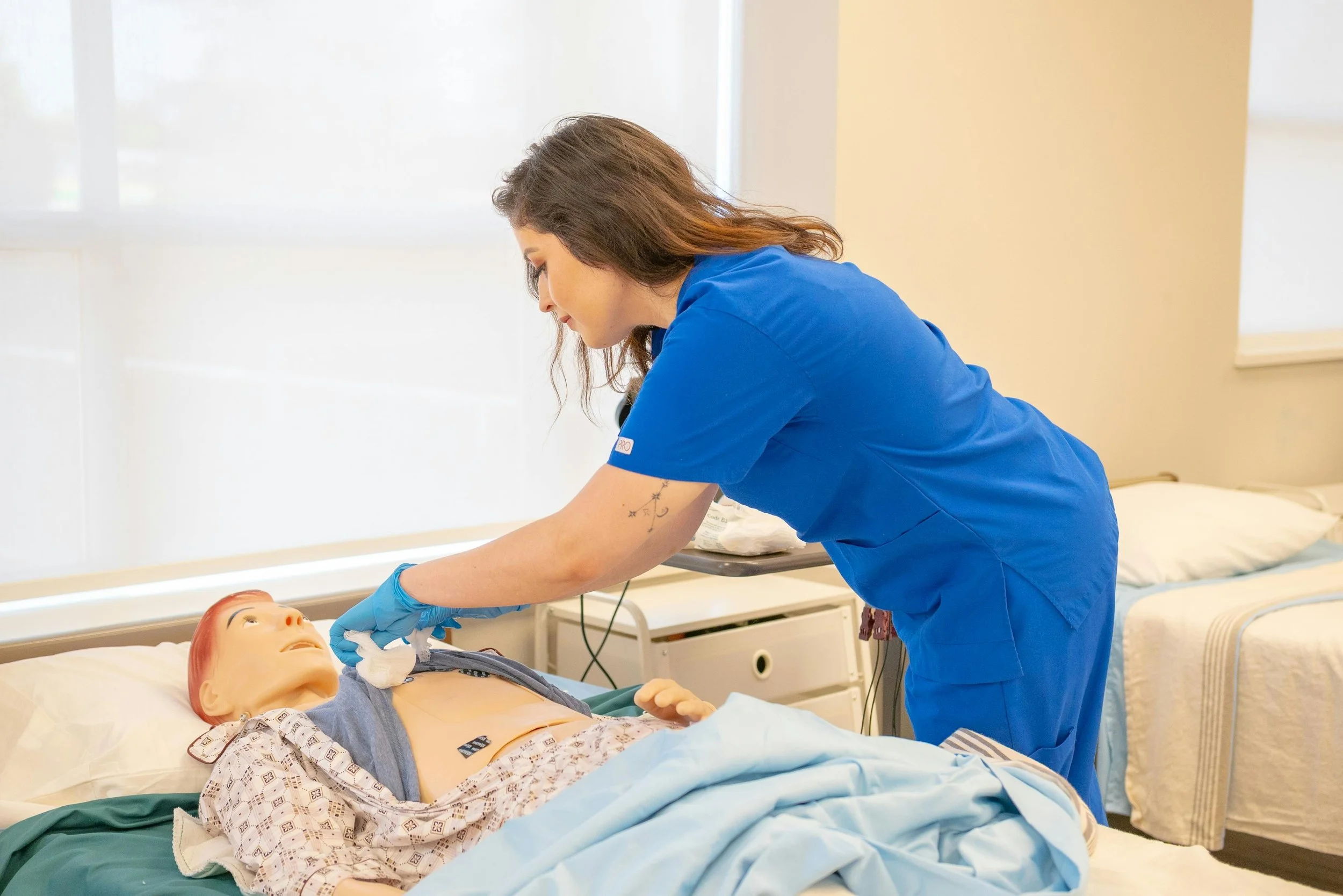A healthcare worker in blue scrubs and gloves is performing a procedure on a medical training mannequin lying in a hospital bed, in a brightly lit room.