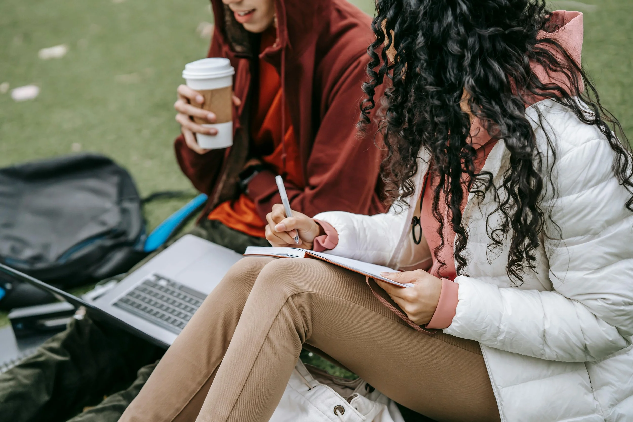 Two young women sitting outdoors on the grass with a laptop, notebook, pen, and coffee, engaged in a discussion or studying.