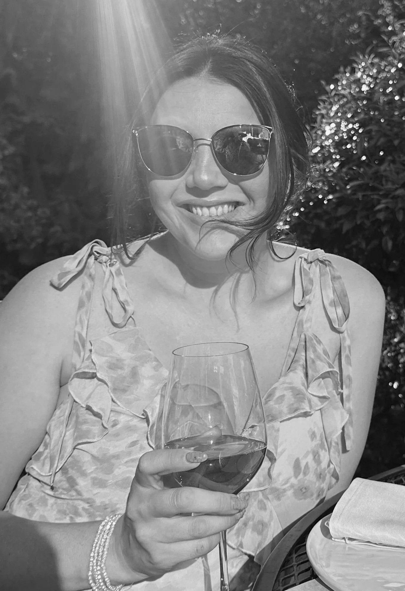 A woman with dark hair wearing sunglasses and a ruffled sleeveless top, smiling while holding a glass of wine outdoors in a sunny setting.