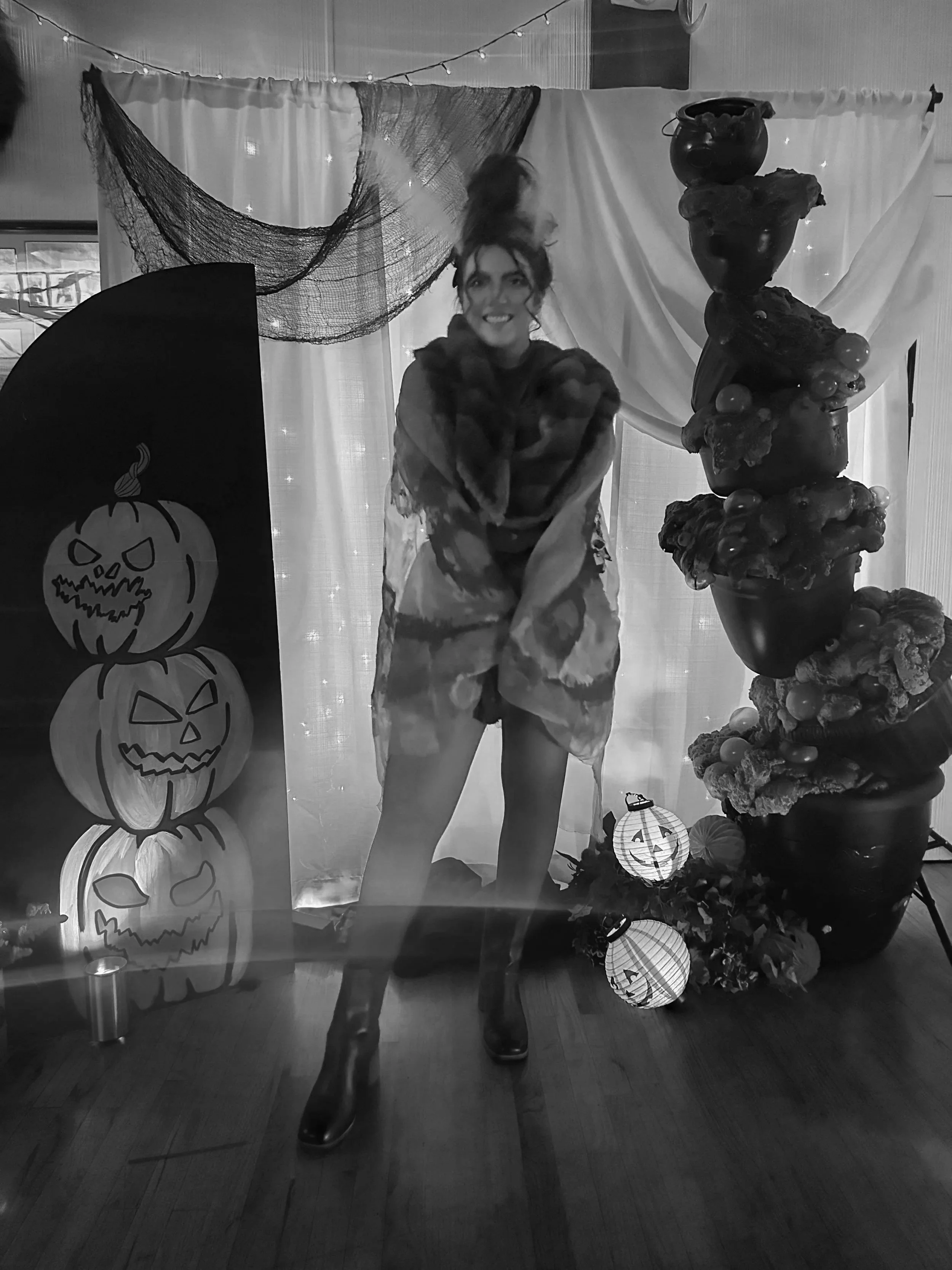 Woman dressed in Halloween costume standing in front of Halloween decorations including jack-o'-lanterns, paper lanterns, and a stacked stone sculpture, with a backdrop of curtains and string lights.