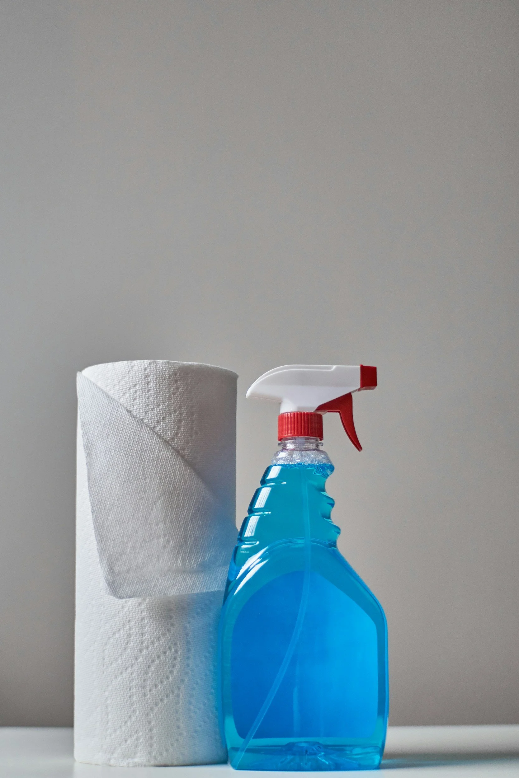 A roll of white paper towels with some of the paper hanging loose, and a spray bottle filled with blue cleaning liquid placed beside it against a plain, light-colored background.