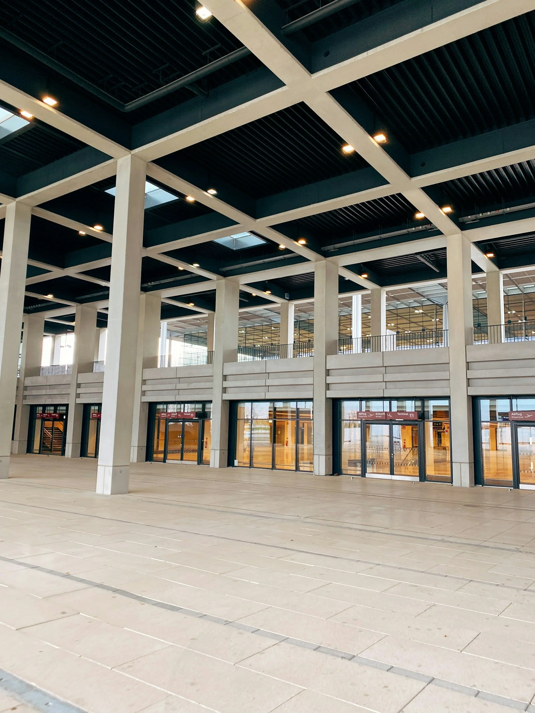 Interior of a modern airport terminal with large open space, multiple flight gates, glass doors, and a balcony area.