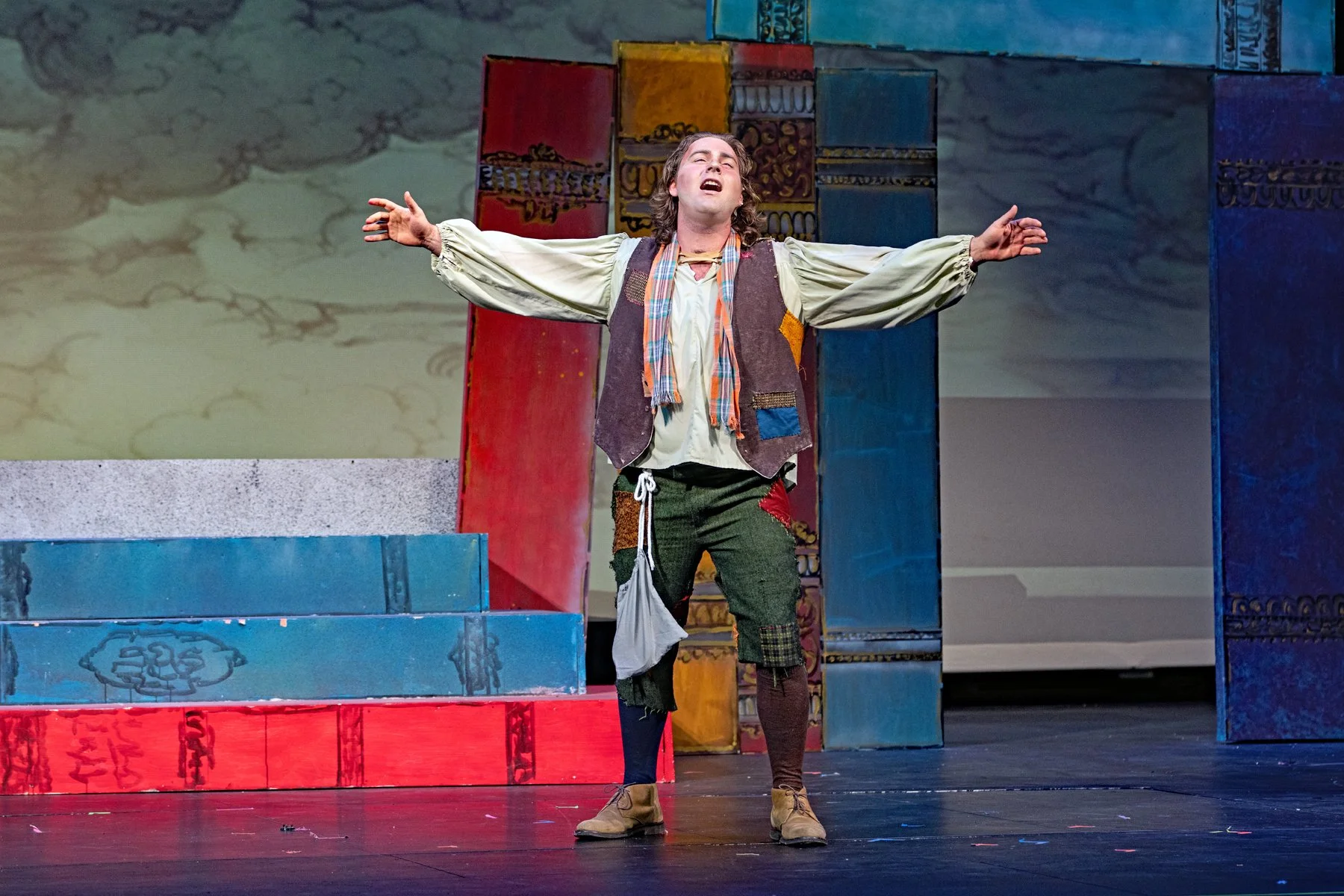 A male actor performing on stage with arms outstretched, wearing a patchwork vest, loose shirt, and patched pants against a colorful, theatrical backdrop.