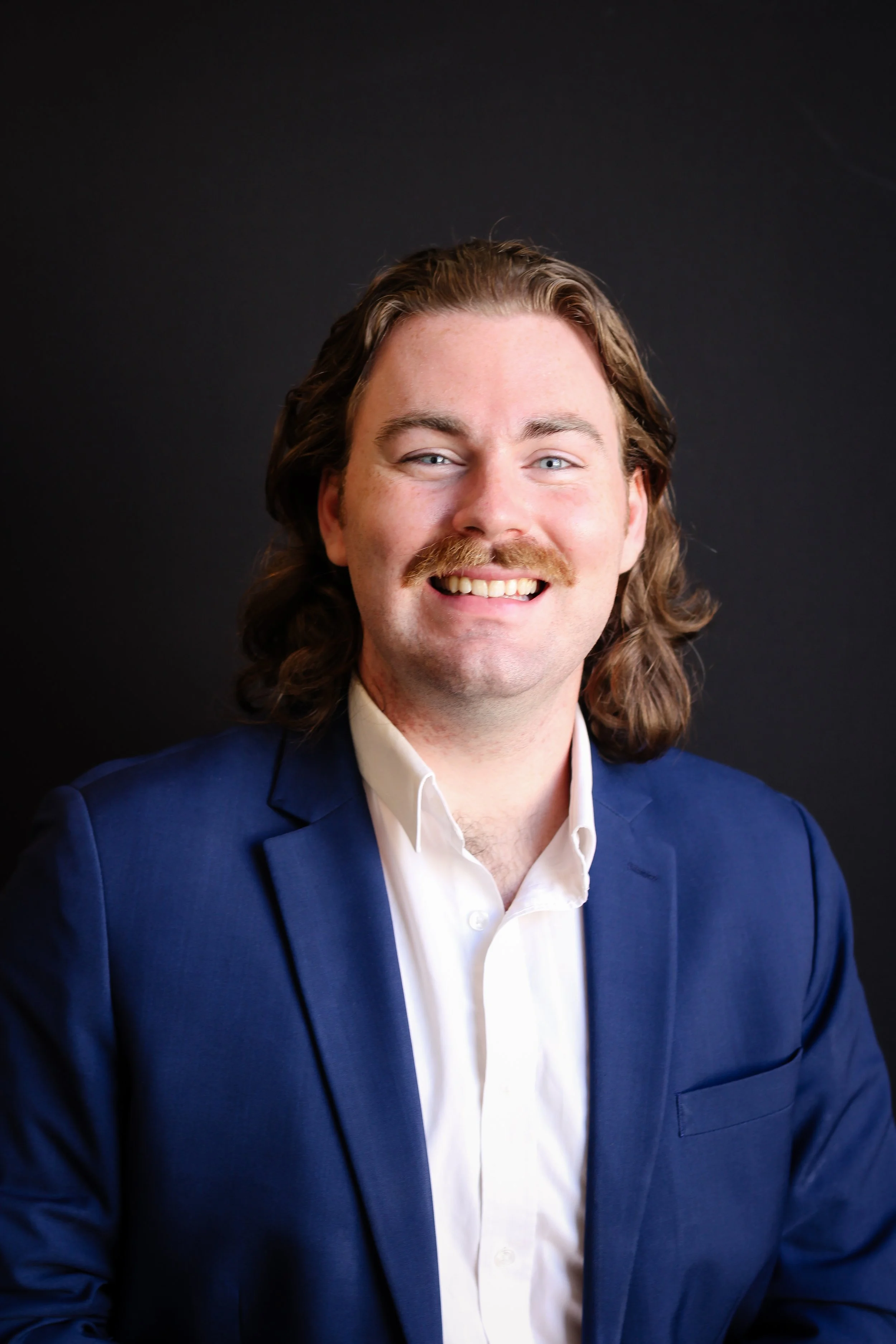 Headshot of a man with shoulder-length wavy brown hair, a mustache, and blue eyes, wearing a navy blue suit and white shirt, smiling against a dark background.