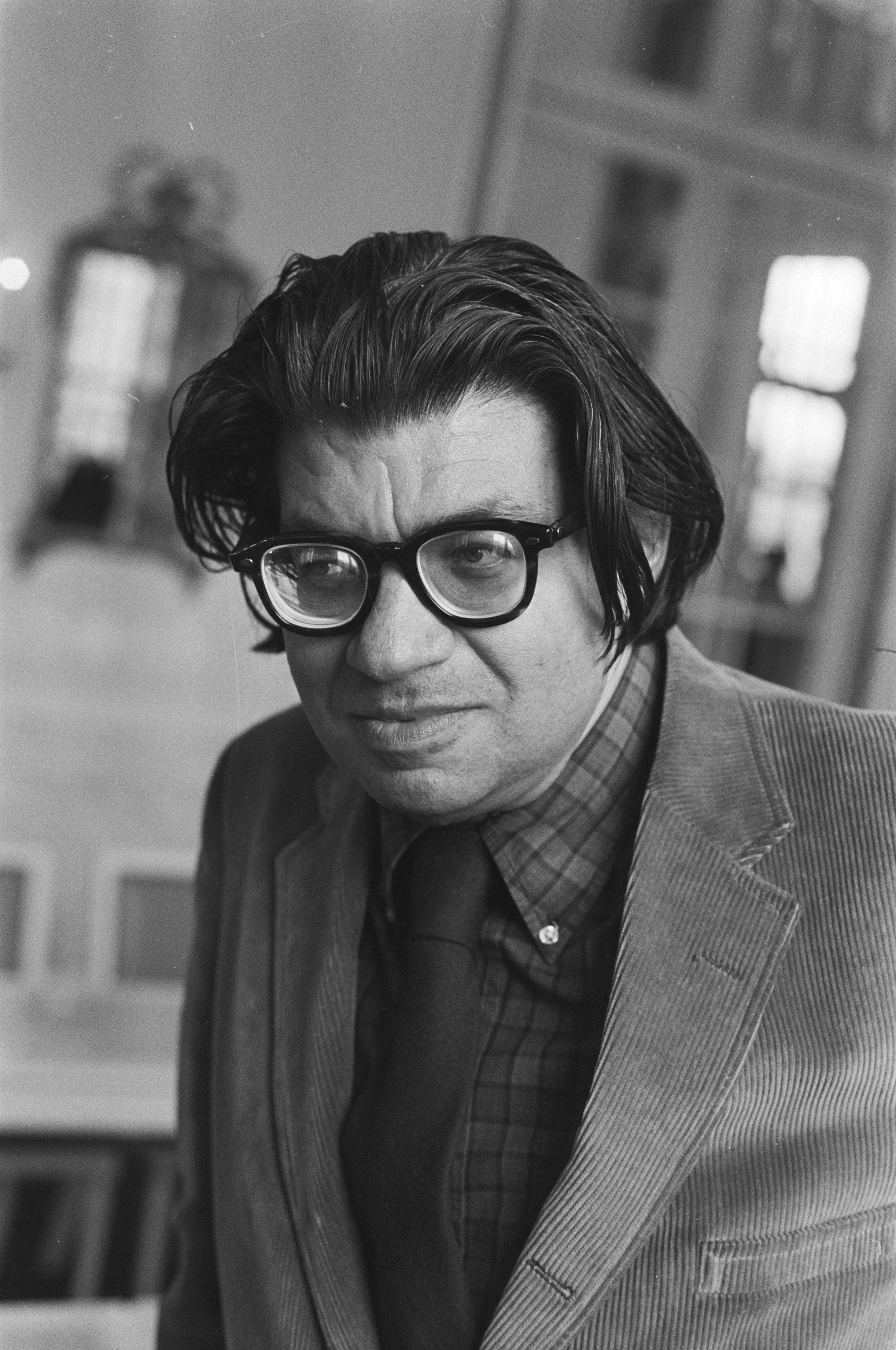 Black and white photo of a man with dark, wavy hair and glasses, wearing a checkered shirt and blazer, looking to the side with a serious expression. Blurred interior background.