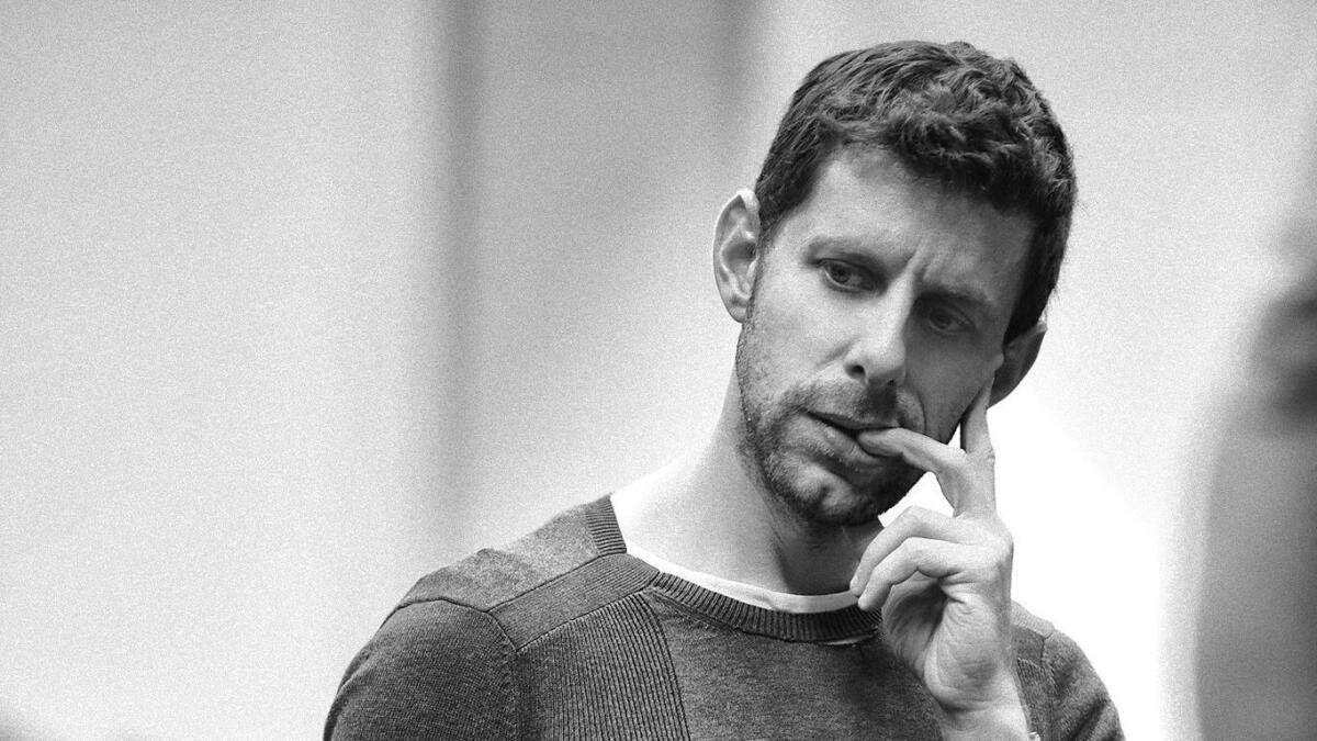 Black and white photo of a man with short dark hair and stubble, resting his chin on his hand with a thoughtful expression.