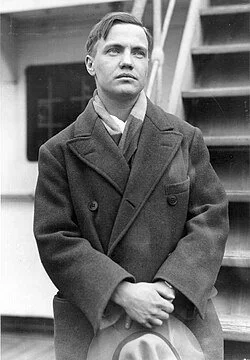 A black and white photo of a man in a dark coat with lapels, standing outdoors near a staircase, looking serious.