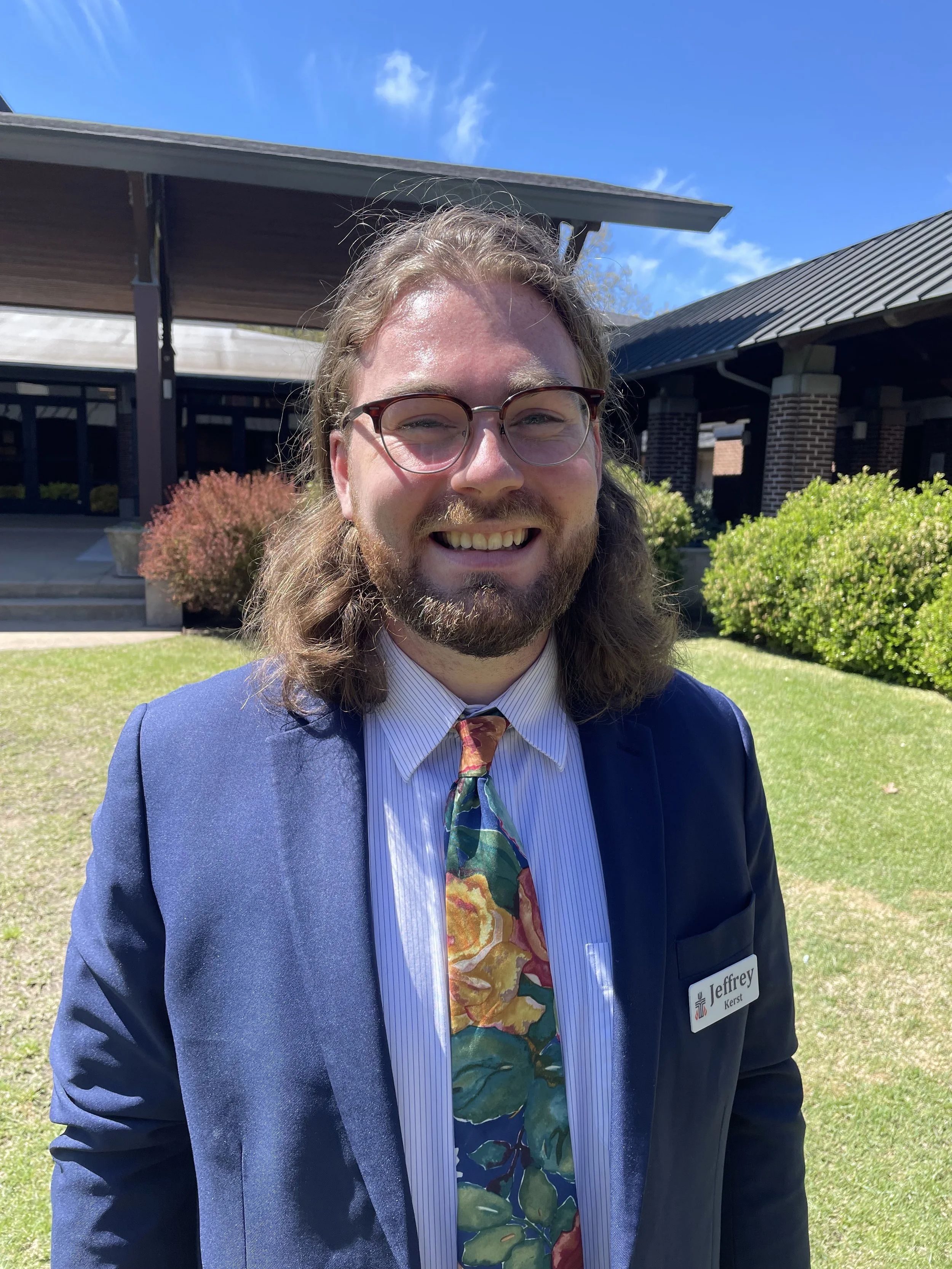 A man with long hair, a beard, and glasses smiling outdoors on a sunny day, wearing a dark suit, a colorful tie, and a name badge that says Jeffrey.