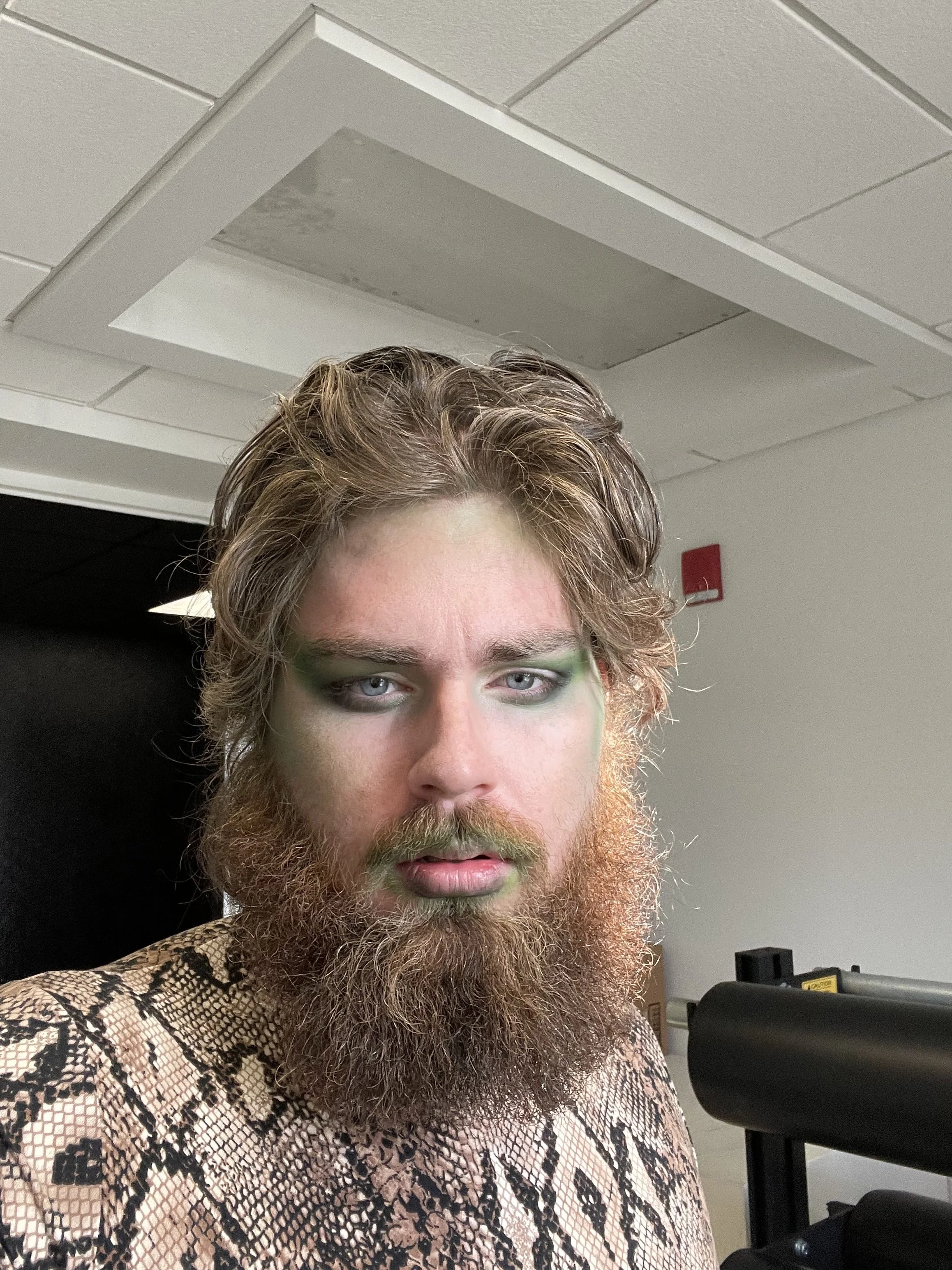 Close-up of a person with light blue eyes, green eyeshadow, brown wavy hair, and a long beard wearing a snakeskin-patterned top. The background shows a white ceiling with a vent and part of a black piece of equipment.