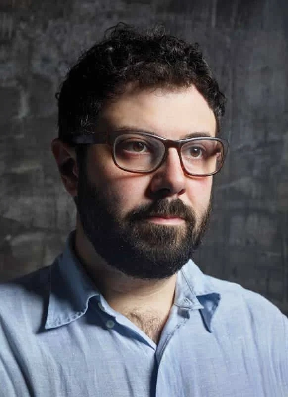 A man with dark curly hair, glasses, and a beard wearing a light blue shirt against a dark textured background.
