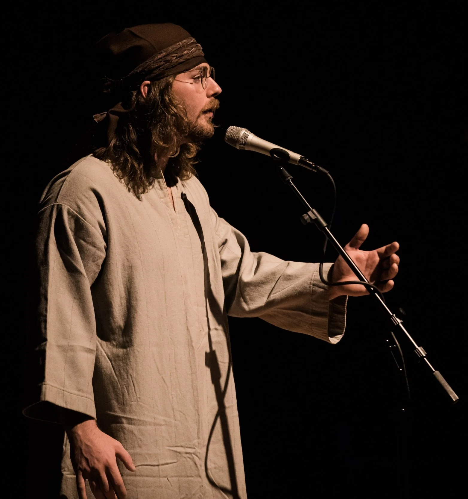 A man with long hair, beard, glasses, and a head covering is performing into a microphone on stage with a dark background.