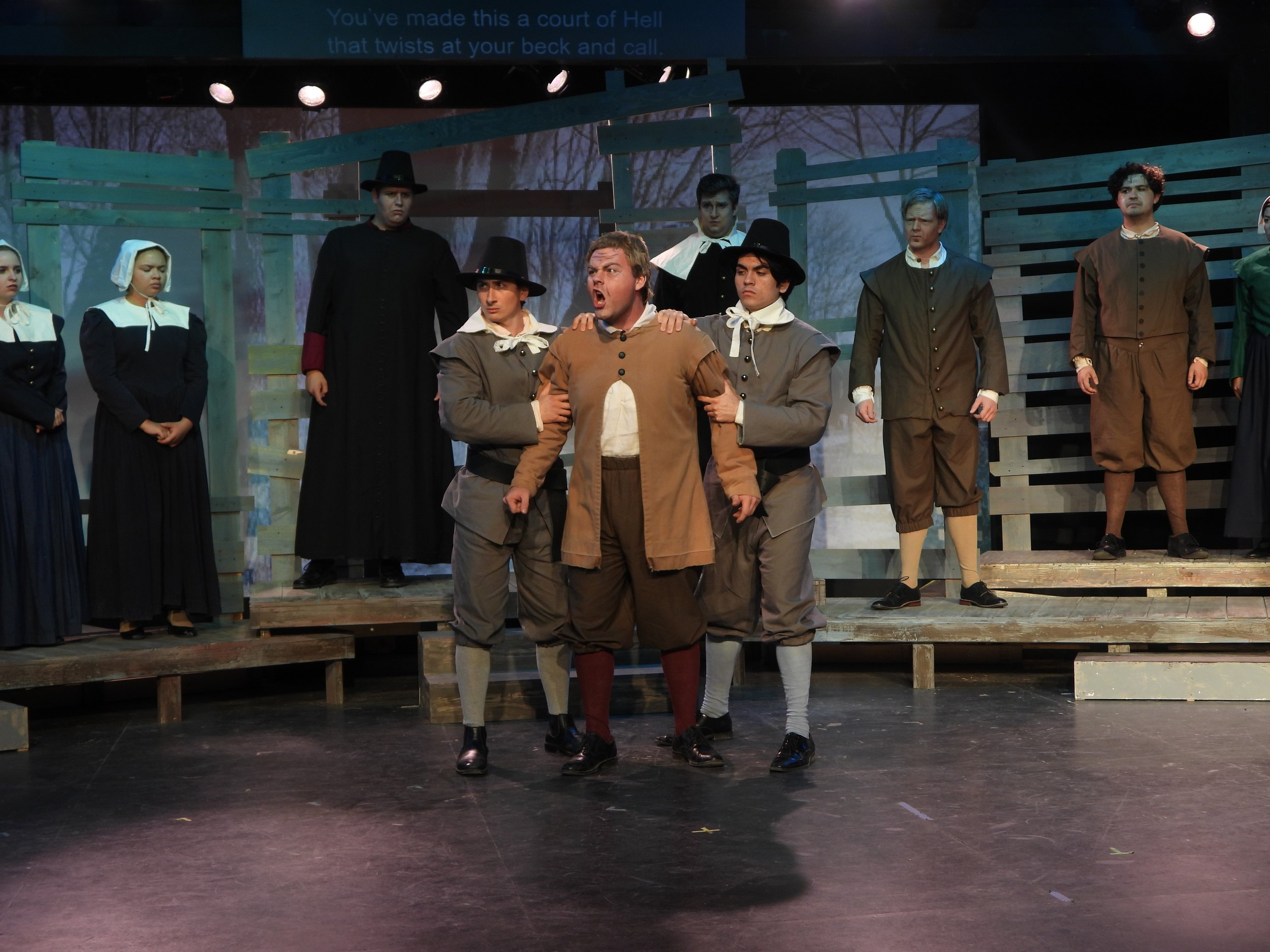 The image depicts a scene from a theatrical play with actors dressed in period costumes on stage. Most characters are wearing old-fashioned clothing, some with bonnets and others with wide-brimmed hats. There are wooden fencing structures in the background, indicating an outdoor or rustic setting. One actor at the center appears to be shouting or speaking passionately, while others show serious or concerned expressions. The background includes a forest scene projected or painted behind the actors.