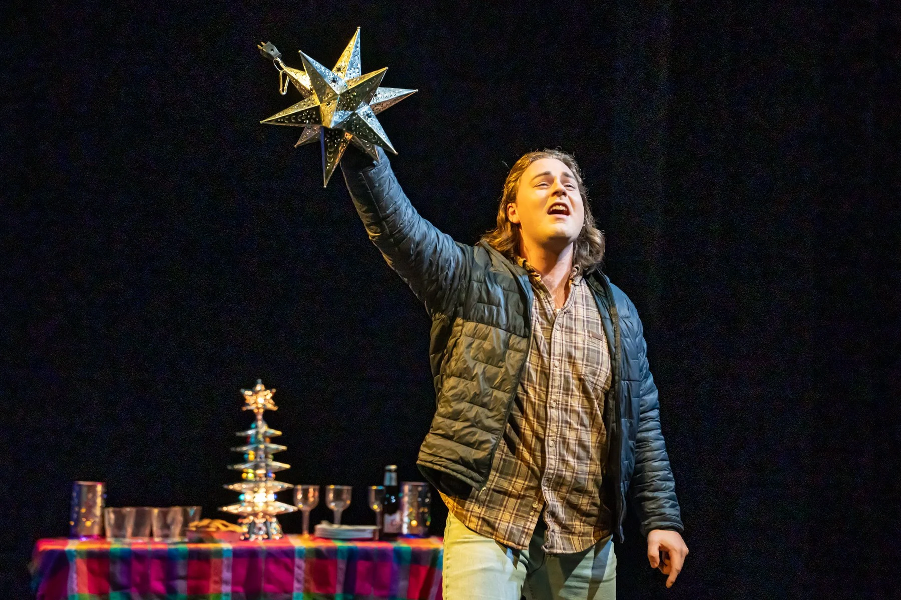 A man with long hair, wearing a plaid shirt and a black jacket, is holding a star-shaped lantern in the air and appears to be speaking or singing. There is a table with a colorful checkered tablecloth in front of him, with various decorative items including a multi-tiered metal star and small glass containers, all set against a dark background.