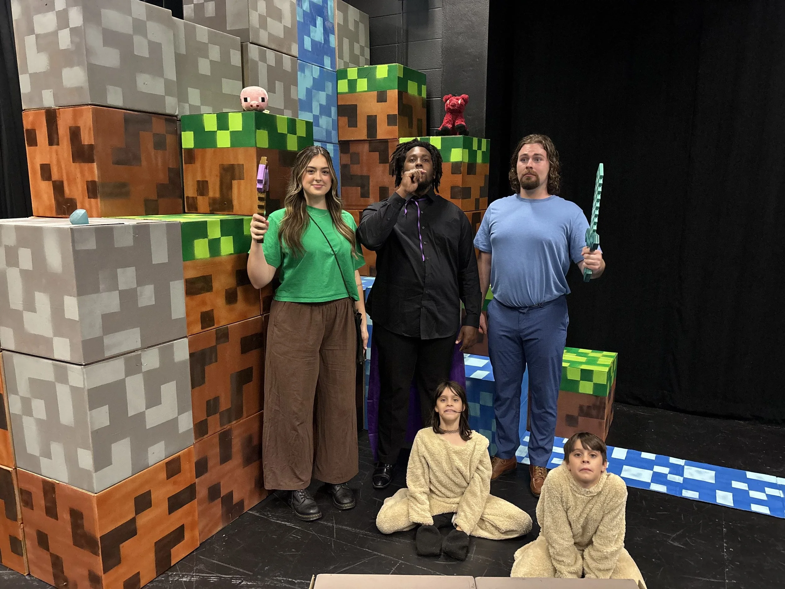 A group of people, including kids, posing in front of a backdrop styled to look like pixelated blocks from Minecraft. The backdrop features large, colorful cubes resembling Minecraft blocks, and some themed stuffed toys are placed on top. The children are dressed in yellow fuzzy costumes, with two sitting on the floor and one standing at the side, while two adults stand in the middle.