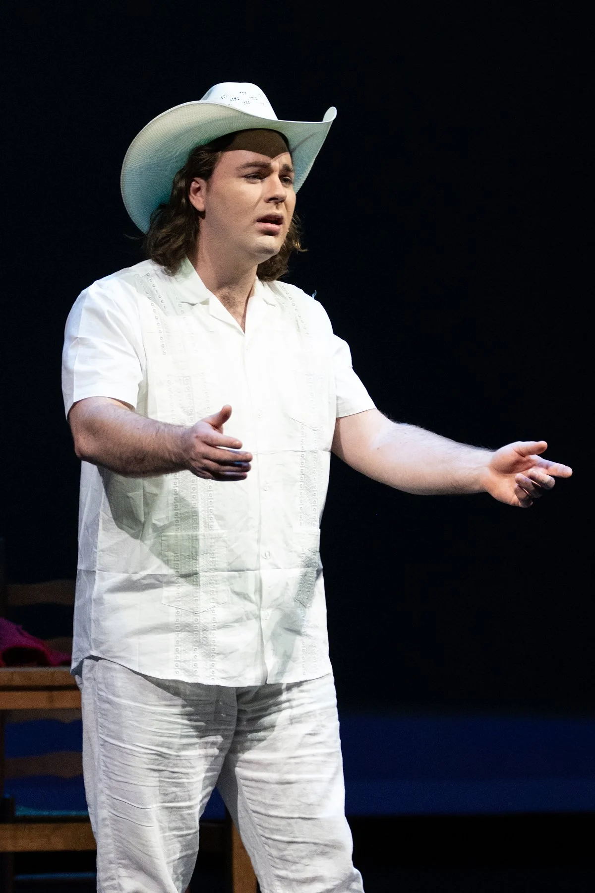 A man dressed in white, wearing a cowboy hat, performing on stage with a black background.