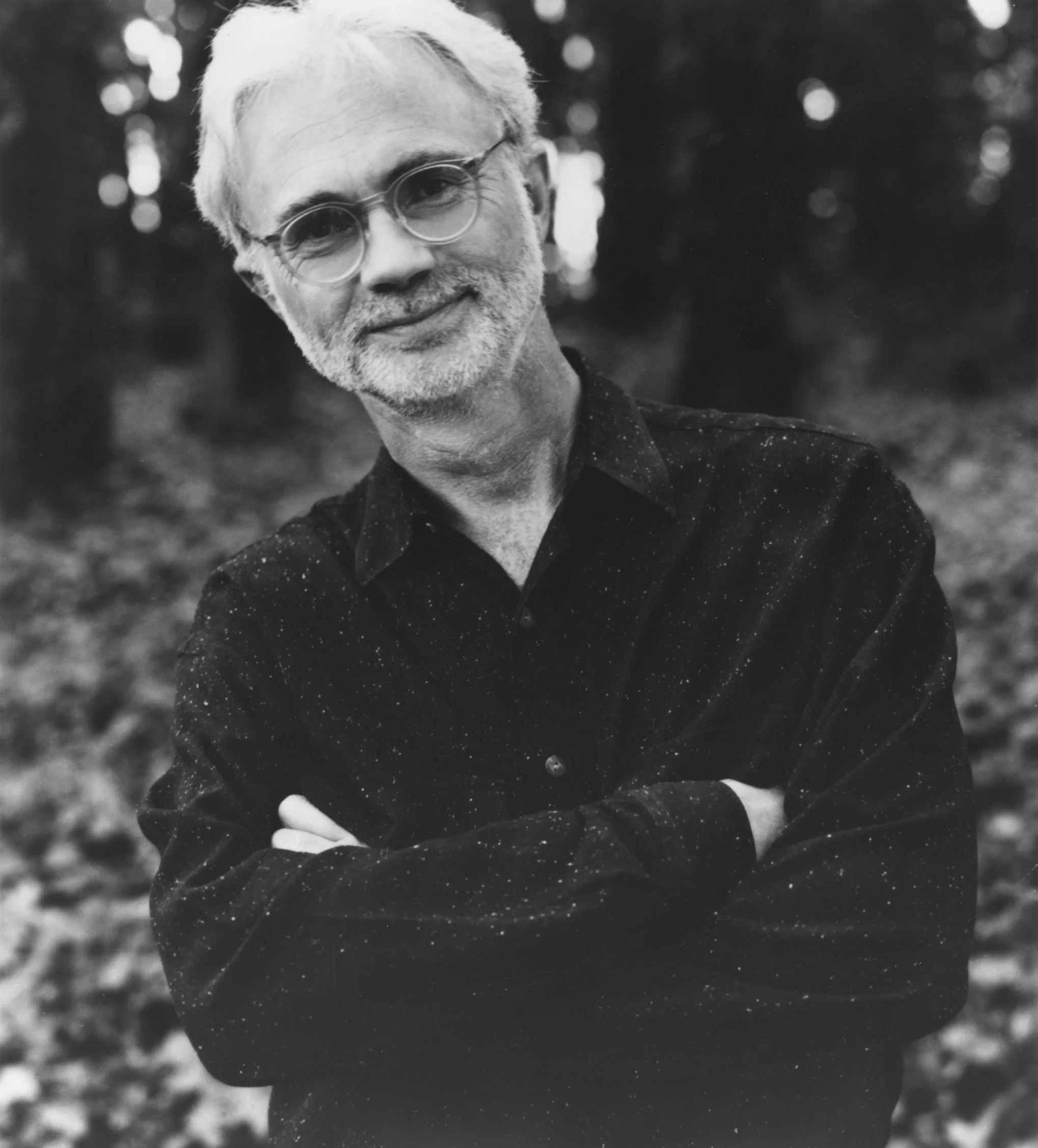 Black and white portrait of a middle-aged man with light hair, glasses, and a beard, wearing a dark, speckled shirt, standing outdoors with blurred trees in the background.