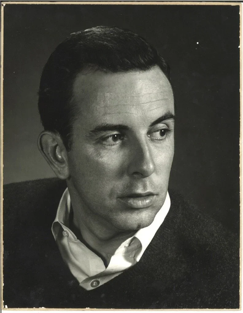 Black and white portrait of a man with slicked-back dark hair, wearing a white collared shirt and a dark sweater, looking to the side.