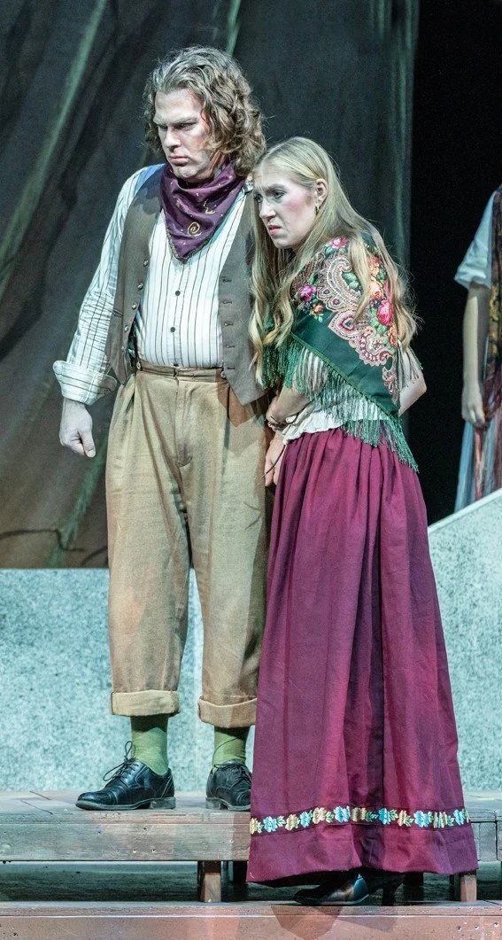 Two actors in period costumes performing on stage, with a wooden platform and a dark, rustic background.