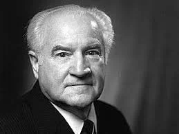 A black and white portrait of an elderly man with white hair and a serious expression, wearing a dark suit and tie.