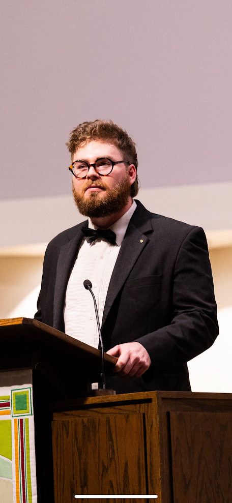A man with a beard and glasses standing at a podium, dressed in a tuxedo with a bow tie, speaking into a microphone in a formal setting.